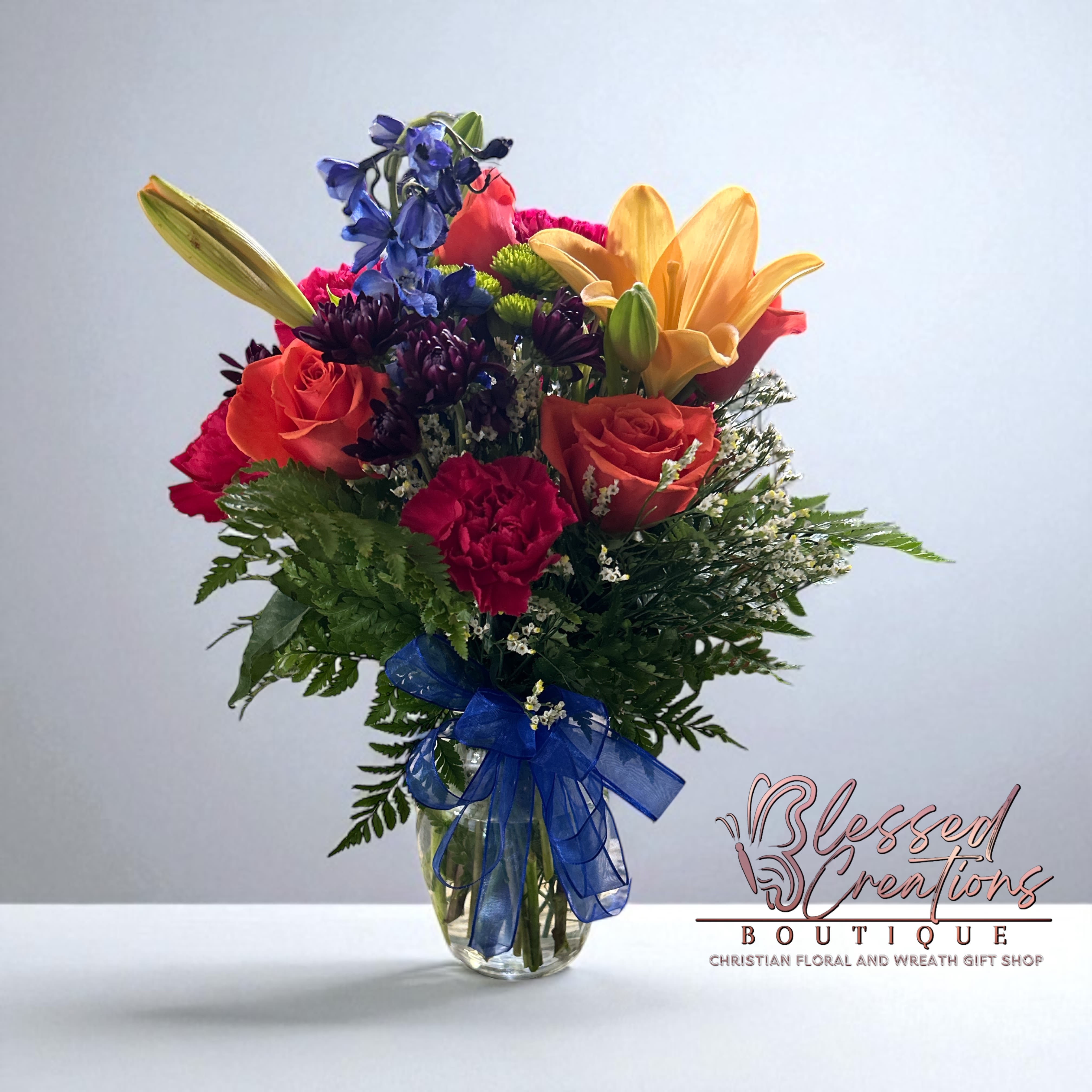 Bright Horizons Bouquet  - Full of color and life, this joyful bouquet features orange roses, golden lilies, red carnations, blue delphinium, purple mums, green button poms, and fresh greenery, finished with a striking blue ribbon bow. Designed in a clear vase, it’s the perfect gift to celebrate birthdays, encouragement, congratulations, or any occasion that calls for a burst of cheer and beauty.