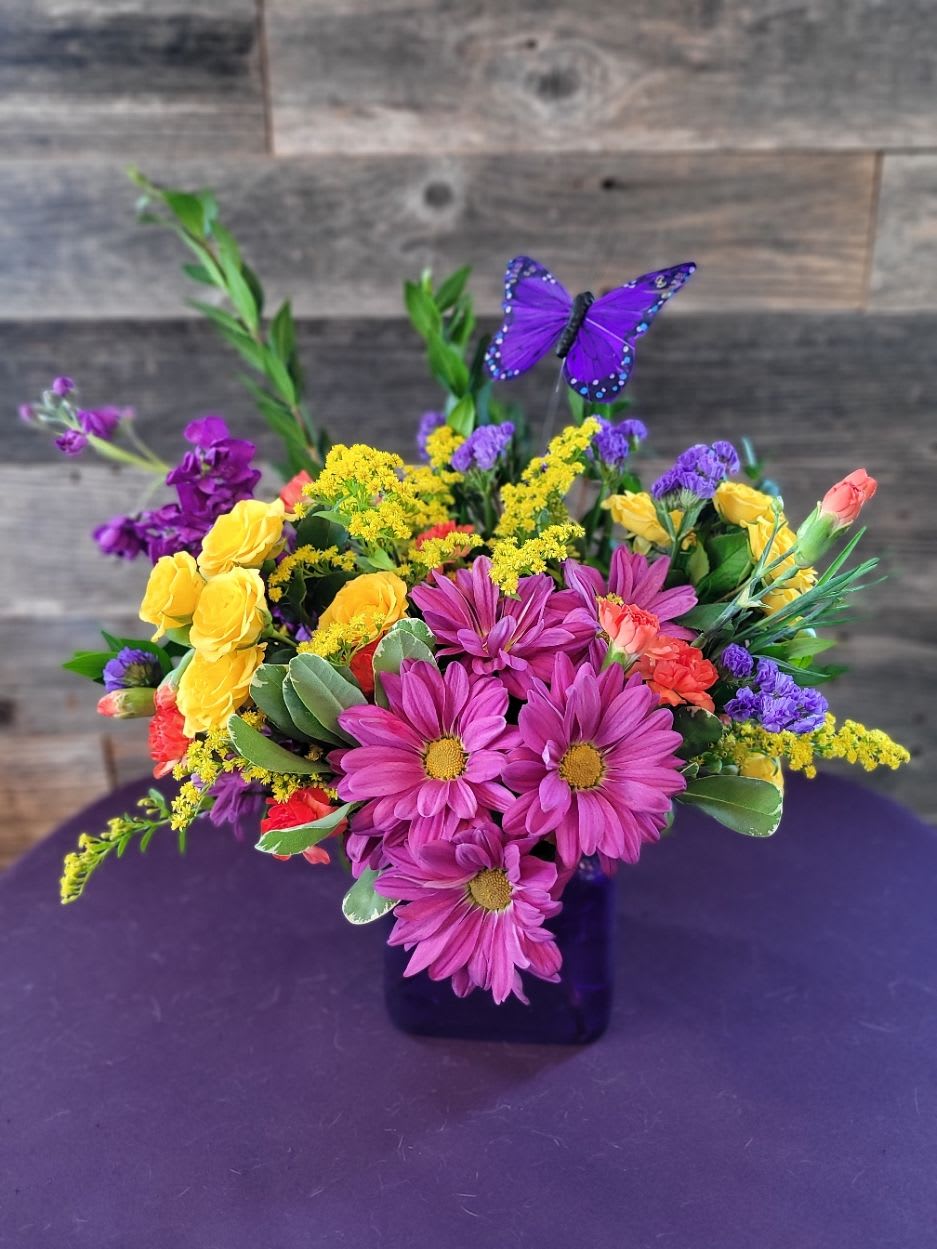 Nature Nature - A kaleidoscope of color and joy! With bright fuchsia , golden yellows, sunny solidago, and pops of coral, and purple come together in a purple cube for a burst of pure happiness. Finished with a charming butterfly, this playful arrangement is perfect for celebrations, thank-yous, or just because.