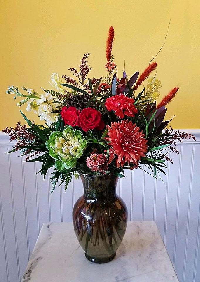 Autumn Opulence - Discover the perfect centerpiece for any sophisticated setting with our vibrant floral arrangement. Featuring lush red spray roses, yellow stock, and exotic orange blooms nestled amidst rich green foliage, all presented in a sleek, dark green and brown ombre glass vase. Ideal for special occasions or to add a touch of elegance to your home.  Order now and bring a burst of color and elegance to your space!  approx dimensions 21 in X 16 in