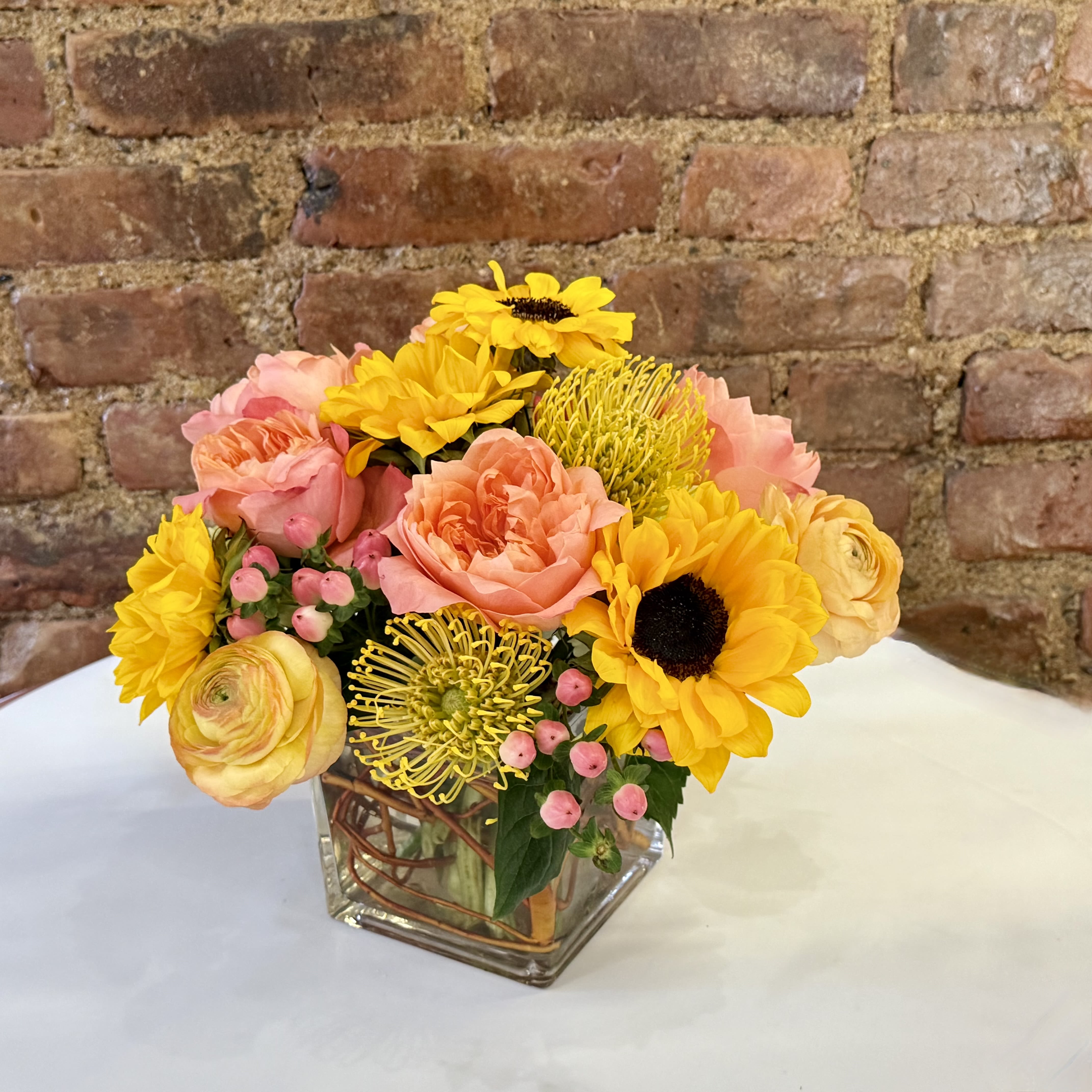 Sunshine Sunshine - Looking to brighten up someone's day? The Sunshine bouquet is the perfect choice! This stunning arrangement features a mix of peach roses, yellow ranunculus, vibrant sunflowers, and yellow pin cushion proteas, all beautifully designed in a modern glass cube vase. Order now to bring a little sunshine into someone's life!