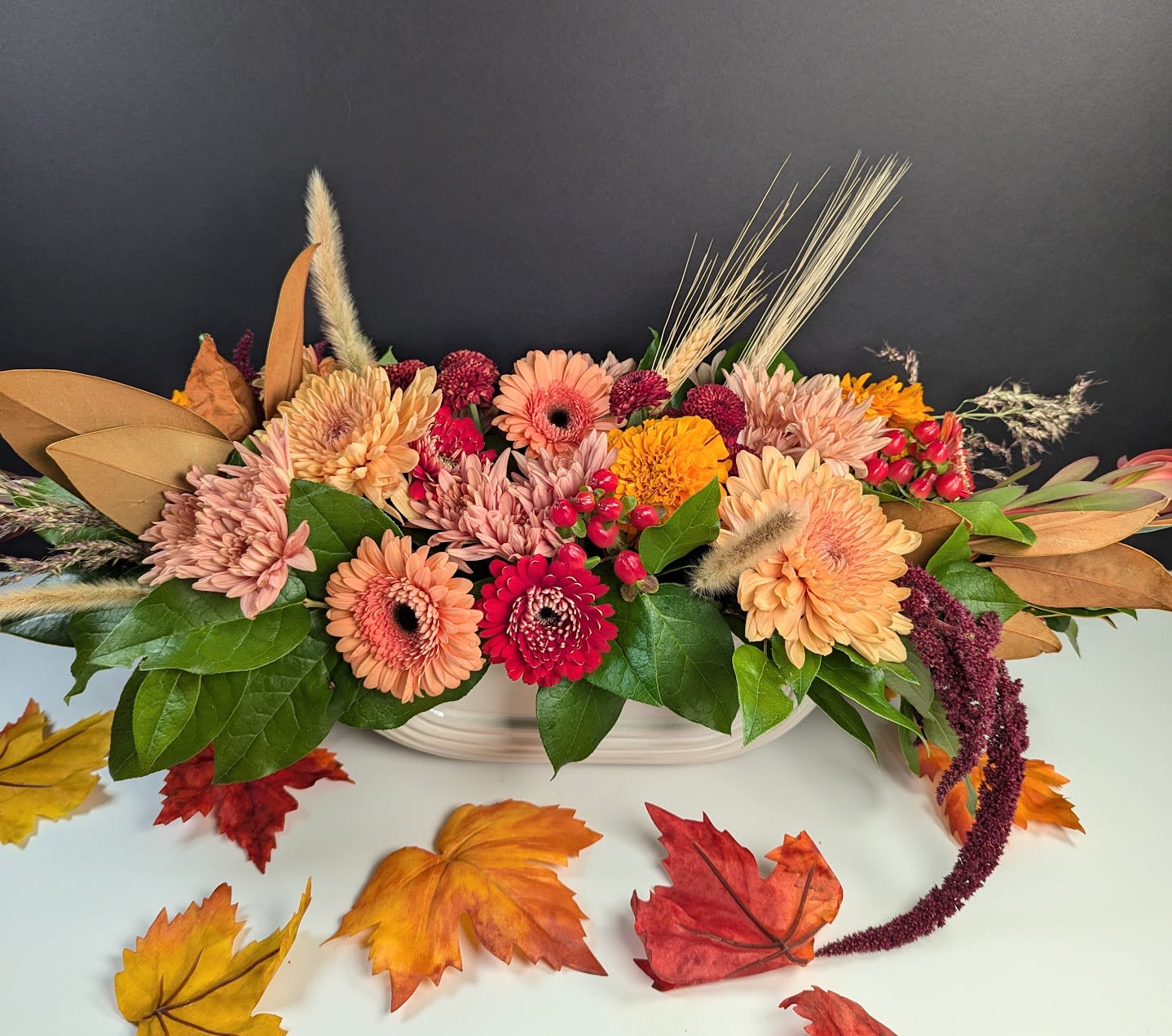 Harvest Moon Centerpiece - Celebrate the rustic charm of autumn with our Harvest Moon Centerpiece. This beautifully crafted arrangement captures the essence of a bountiful harvest, featuring:  -Light orange mums -Red and peachy gerberas -Burgundy pompoms -Burgundy amaranthus -Wheat -Millet -Marigolds -Hypericum berries  Each element in this centerpiece is thoughtfully arranged to evoke the warmth and richness of the season. The natural textures and deep, earthy tones create a stunning display that is perfect for your fall gatherings. Whether as a dining table centerpiece or a festive decoration, the Harvest Moon Centerpiece brings the beauty of the harvest season into your home, making every occasion feel special.