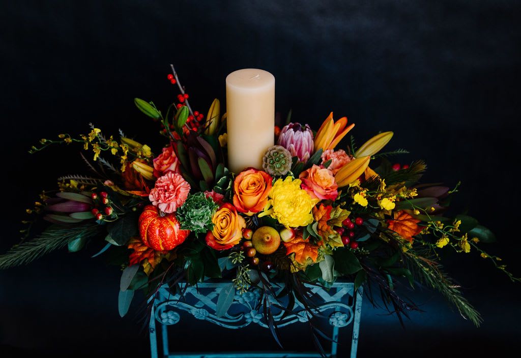 Harvest Glow Centerpiece Harvest Glow Centerpiece - Warm up your autumn table with the Harvest Glow Centerpiece—a lush arrangement of fiery orange roses, golden lilies, protea, berries, and fall foliage, all surrounding a soft pillar candle. Perfect for Thanksgiving gatherings or cozy seasonal decor, this piece brings the beauty of the harvest season indoors with elegance and charm.