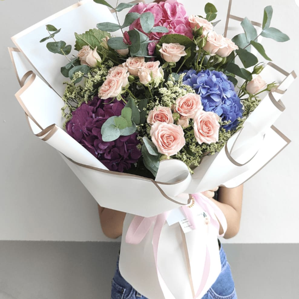 Color Me Happy - Celebrate life with this vibrant bouquet — a joyful harmony of color and texture. Featuring lush hydrangeas in vivid pink, royal purple, and sky blue, accented by delicate blush spray roses, seeded greenery, and touches of eucalyptus, this arrangement is an ode to beauty in full bloom.  Wrapped in crisp white paper with rose-gold edges and finished with a soft pink ribbon, it's the perfect gift for birthdays, milestones, or simply to say “you matter.”  A burst of elegance for the heart that blooms with joy.