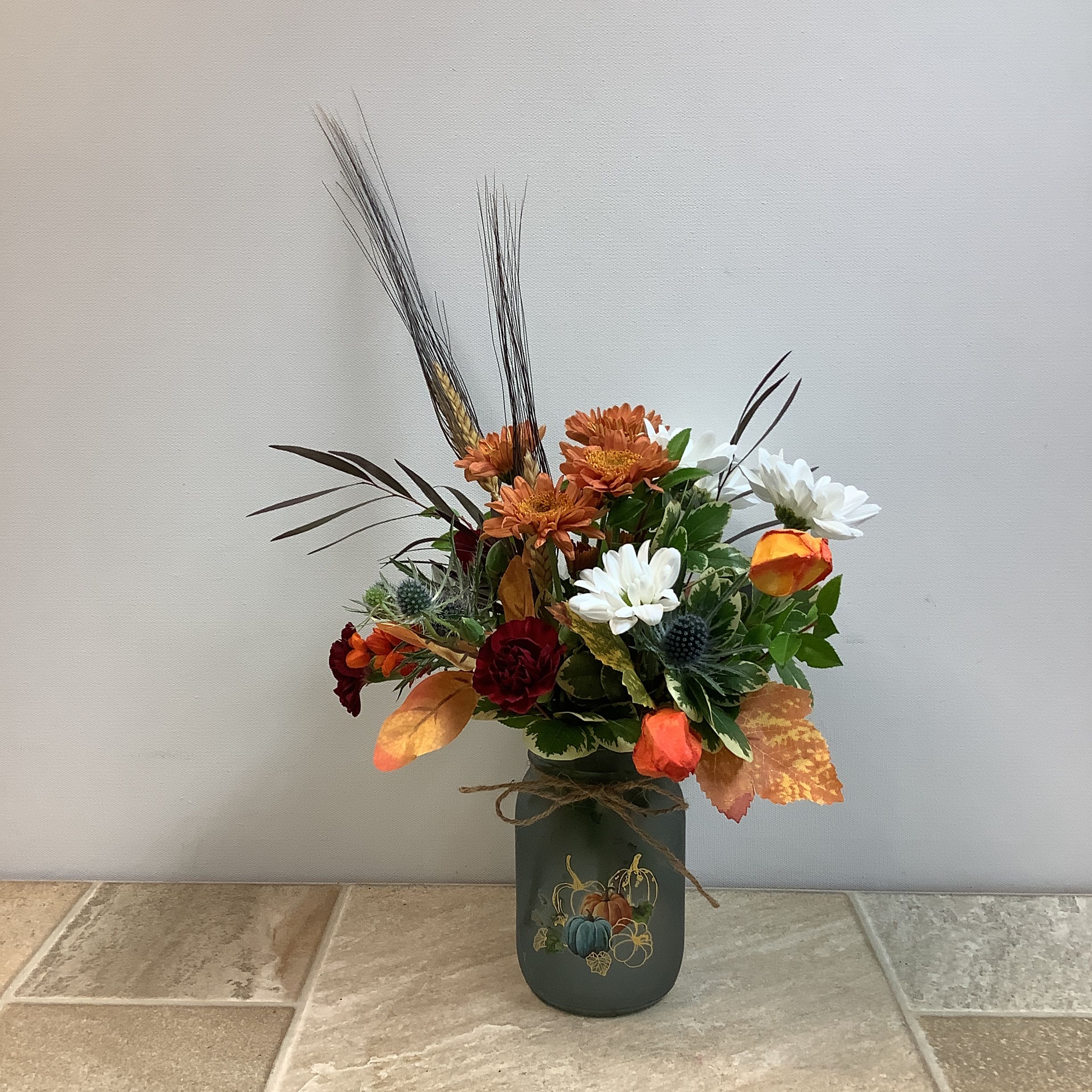 Little Pumpkin Jar Little Pumpkin Jar - This arrangement has a warm, seasonal charm with an autumn harvest theme. The bouquet features burnt orange mums, deep burgundy blooms, cheerful white daisies, thistle accents, and fall foliage in shades of gold and orange. Tall wheat stalks and rustic greenery add texture and height, while the painted vase with pumpkins and gourds ties the whole look to the harvest season. A twine bow around the vase gives it a country, homespun feel. It measures 9” wide x 13” tall.