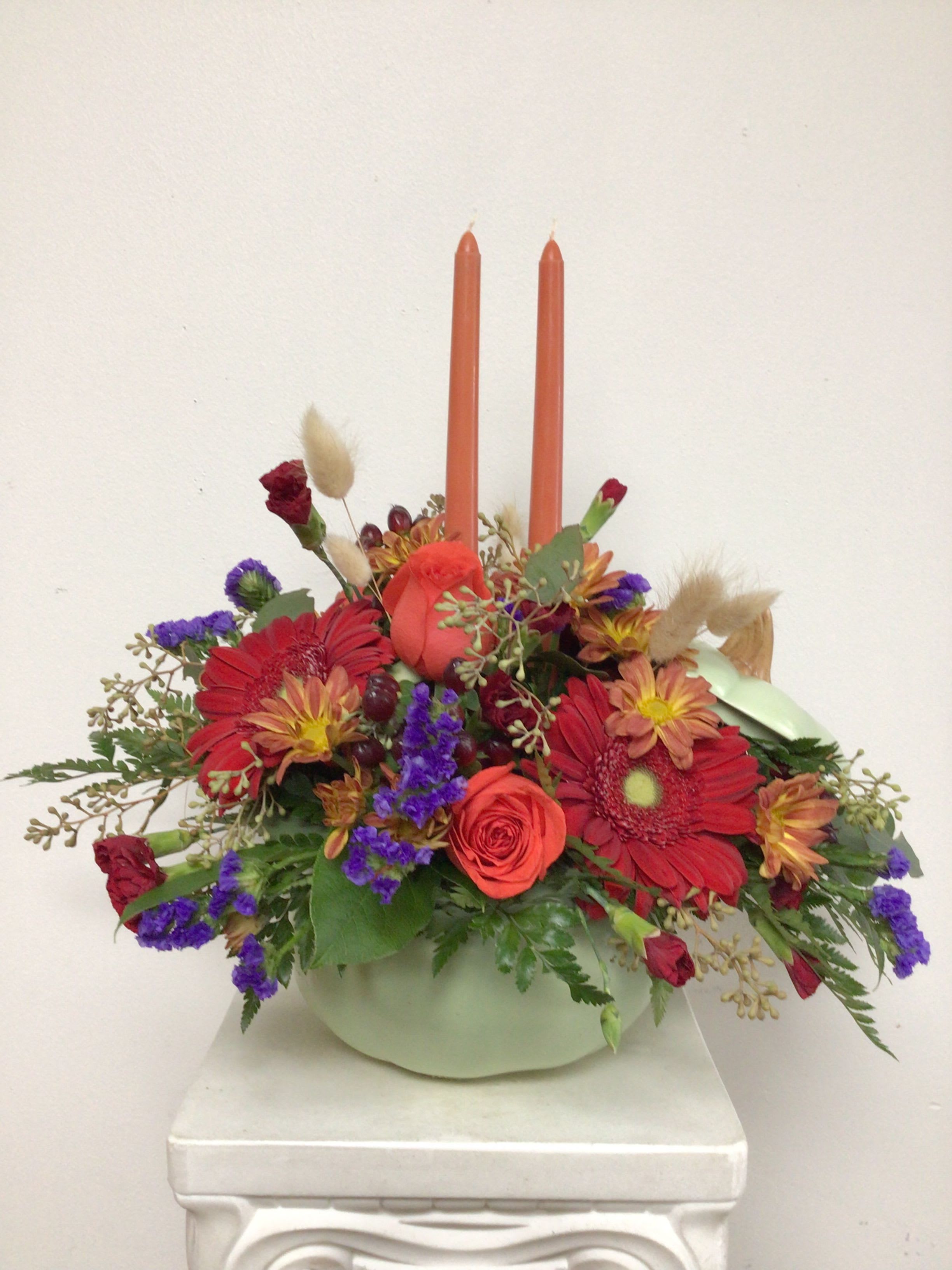 Harvest - Ceramic pumpkin with lid is arranged with red gerbera daisies, orange roses, red miniature carnations, butterscotch daisy mums, purple statice, mahogany hypericum berries, seeded eucalyptus, dried wheat and 2 taper candles. Approximate size is 15 inches wide by  16 inches tall.