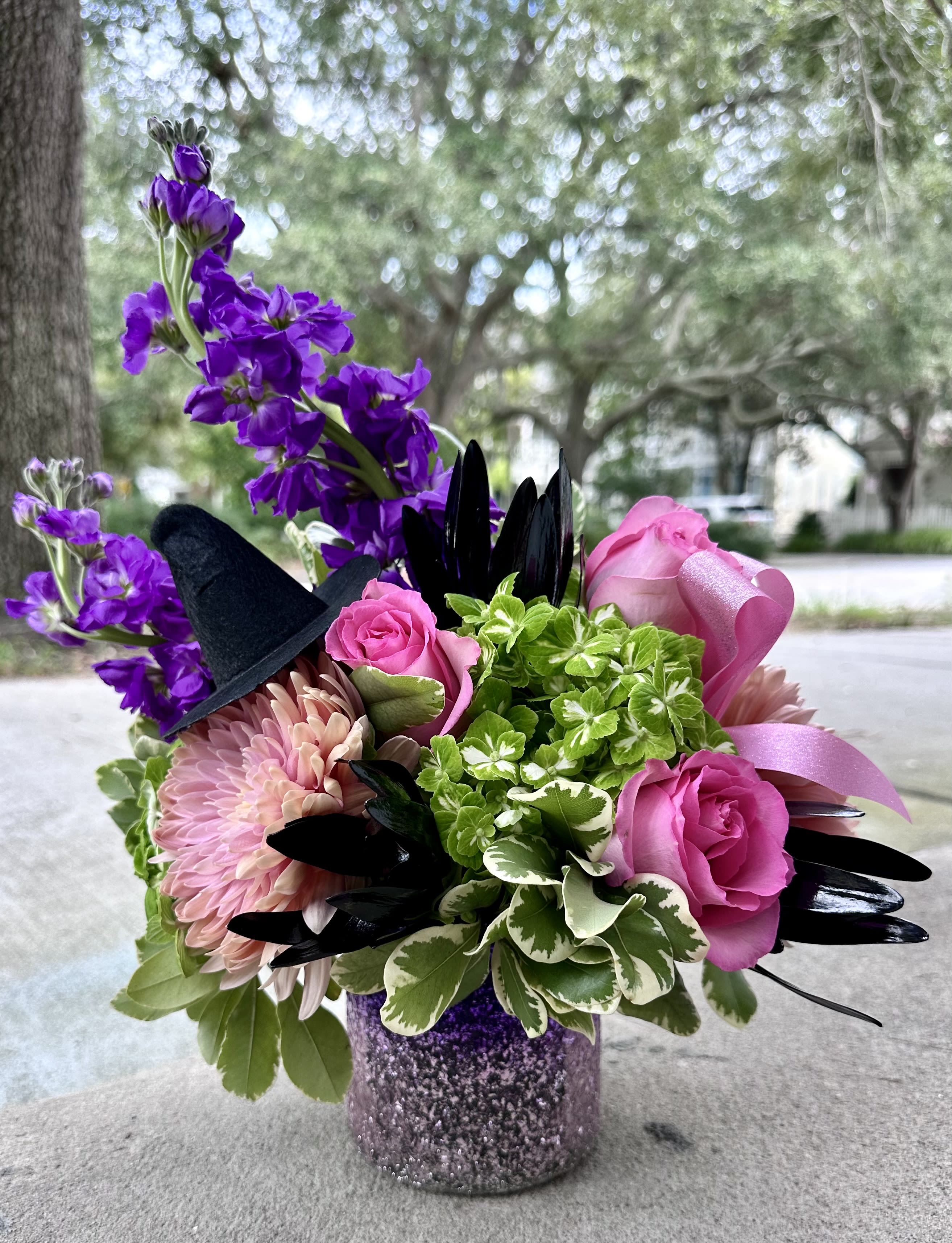 Wicked Blooms - Cast a spell this season with our enchanting Wicked Blooms arrangement. Inspired by the POP-ULAR movie, this arrangement shows bold purple stock and vibrant pink roses that meets striking pink chrysanthemums and fresh green hydrangea, all accented with dramatic black foliage and a witches hat and sparkly pink ribbon. A little big magical, a little bit mischievous - the perfect centerpiece for a wickedly stylish fall *Picture is a deluxe size*