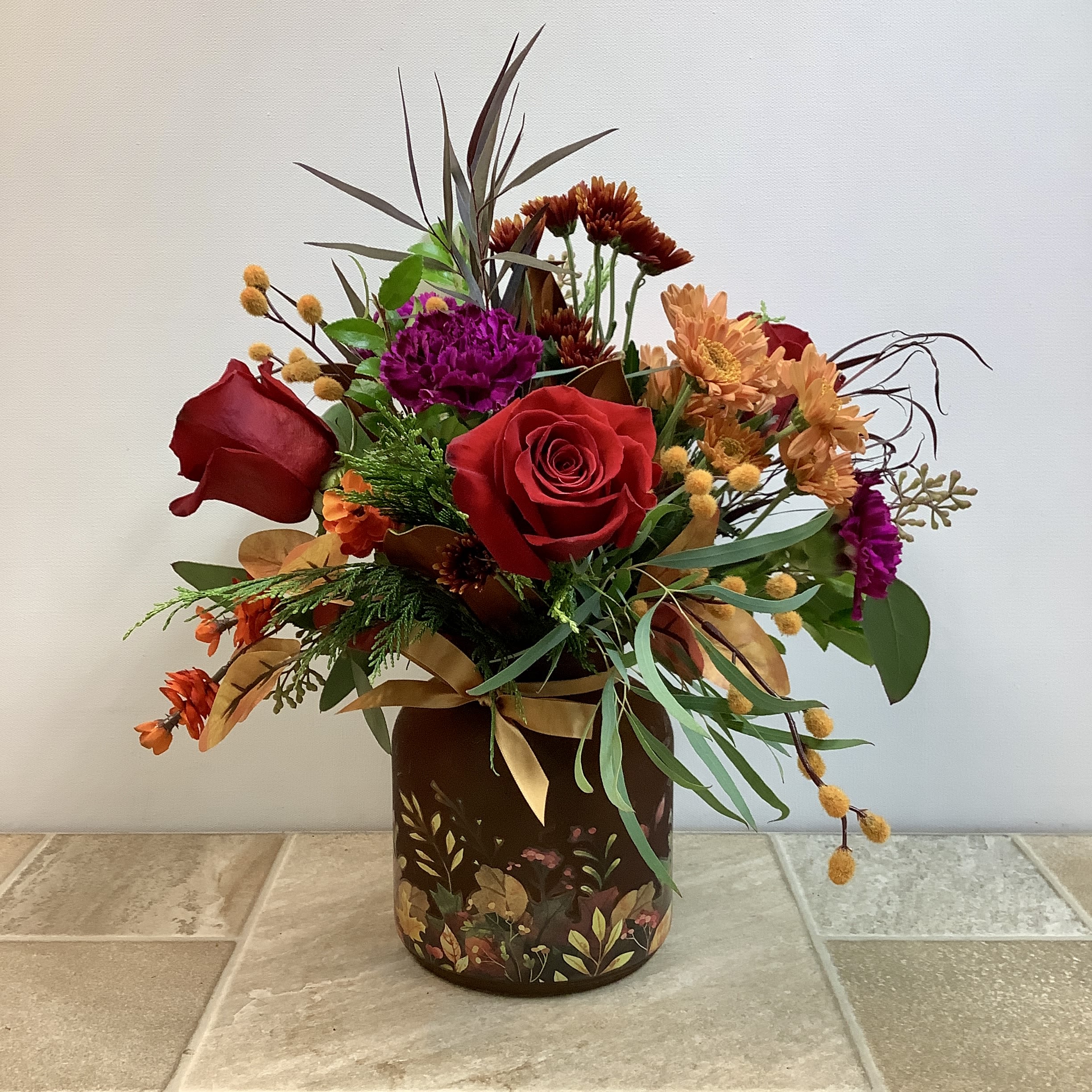 Crimson and Gold Crimson and Gold - This bouquet features a warm and rich autumn palette with bold red roses as the focal point, accented by deep magenta carnations, orange mums, and small orange blooms. Wispy brown foliage and eucalyptus give it movement, while the golden ribbon ties everything together. The arrangement is displayed in a vase adorned with floral details, enhancing its seasonal charm. It measures 14” wide x 17” tall. Primary flowers: rose, carnation, mum