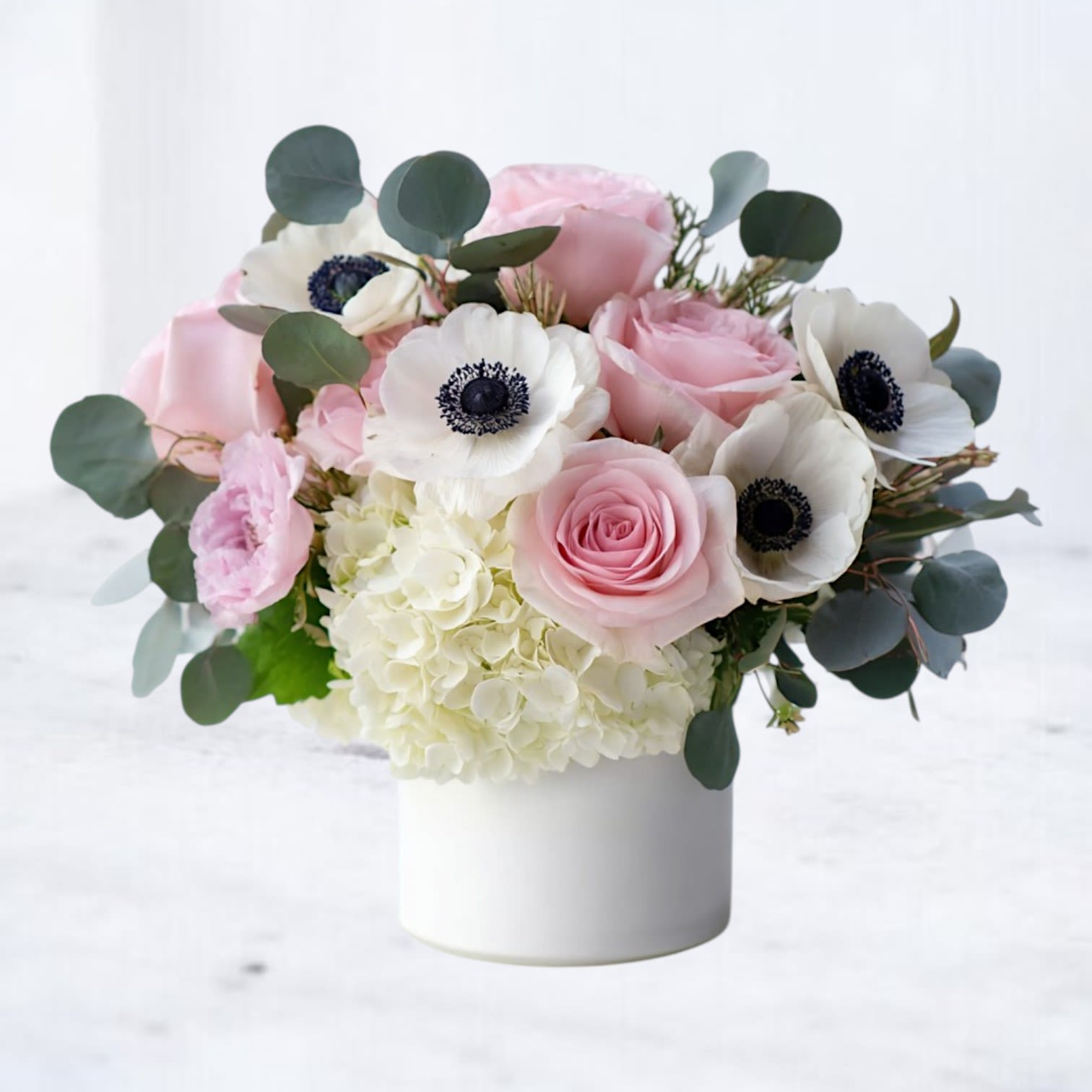 Anemone Alchemy - Soft yet striking, Anemone Alchemy is a luxurious floral arrangement that captures the magic of transformation through nature’s most enchanting elements. Featuring delicate pink roses, pristine white anemones with their captivating black centers, fluffy white hydrangea, and lush eucalyptus, this bouquet evokes a sense of calm wonder.  Designed in a modern white ceramic vase, it’s perfect for celebrating life’s most tender moments—whether you’re sending love, offering comfort, or simply brightening someone’s day with an elegant gesture.