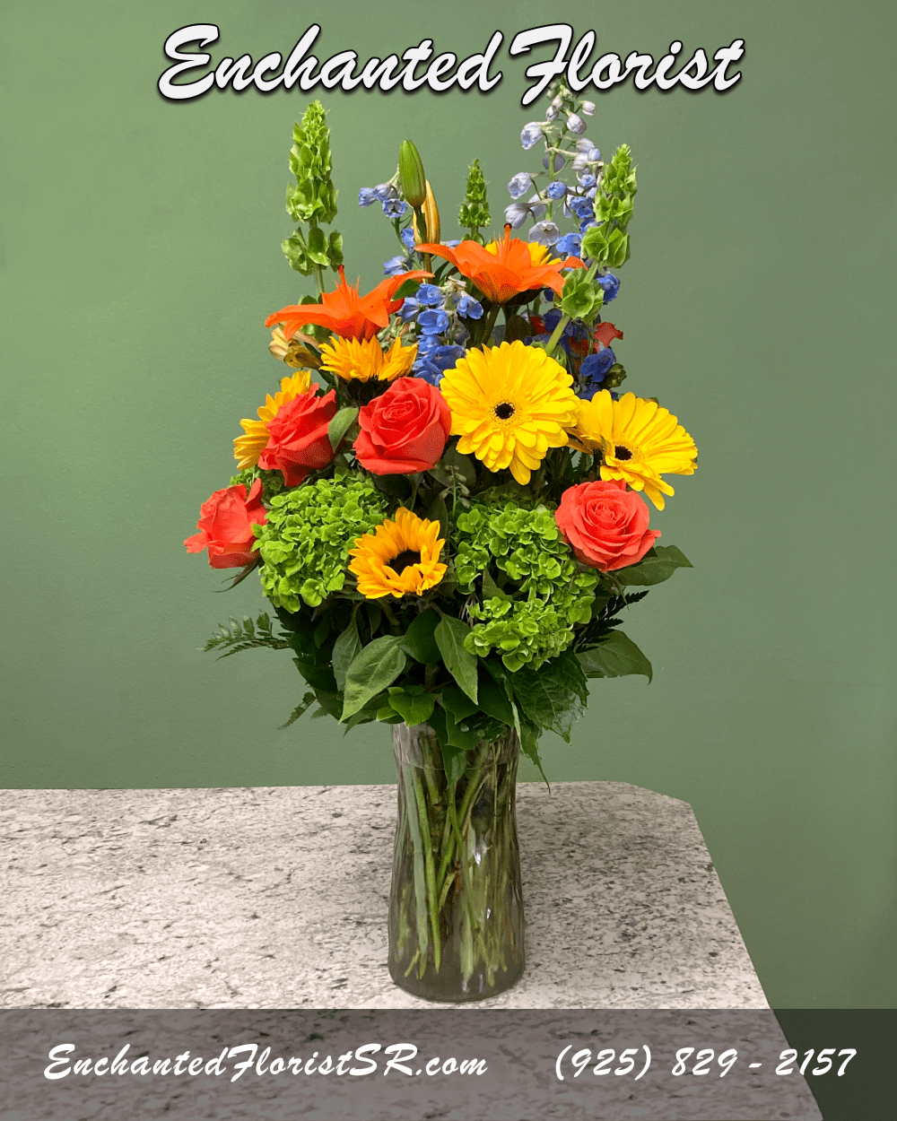Radiant Joy - Mixed Arrangement - A tall and vibrant mixed arrangement showcasing bright, cheerful shades of orange, yellow, and blue. Featuring an array of premium blooms including roses, sunflowers, 'tiger' lilies, delphinium, hydrangeas, and gerbera daises. Accented with 'bells of Ireland', lush lemon leaves, and leather ferns. Presented elegantly in a clear glass cylindrical vase.  This radiant mixed arrangement is perfect for brightening someones day, or adding a pop of color to any room.  Arrives In: Cylindrical clear glass vase  Orientation: Any-Sided  Approximate Dimensions: 28&quot; Tall 14&quot; Wide 6&quot; Vase Diameter