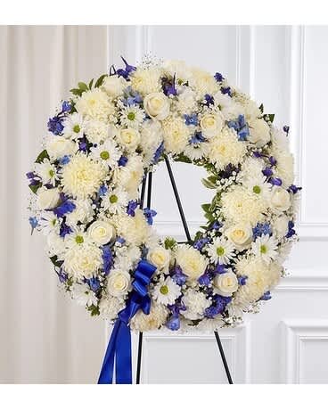 Serene Blessings Standing Wreath- Blue and White - Helping someone find peace after the loss of a loved one can be difficult. If you want to offer your condolences through flowers, our timeless standing wreath in blue and white is a beautiful choice. Crafted with a mix of soothing blooms for a lush, full presentation, this touching tribute helps you express all the love and support you have in your heart when they need it most. One-sided standing wreath arrangement with white roses, football mums, daisy poms and carnations, shades of dark and light blue delphinium; accented with babyâs breath, assorted greenery and blue ribbon Appropriate for the funeral home Our florists hand-design each arrangement, so colors and varieties may vary based on local availability