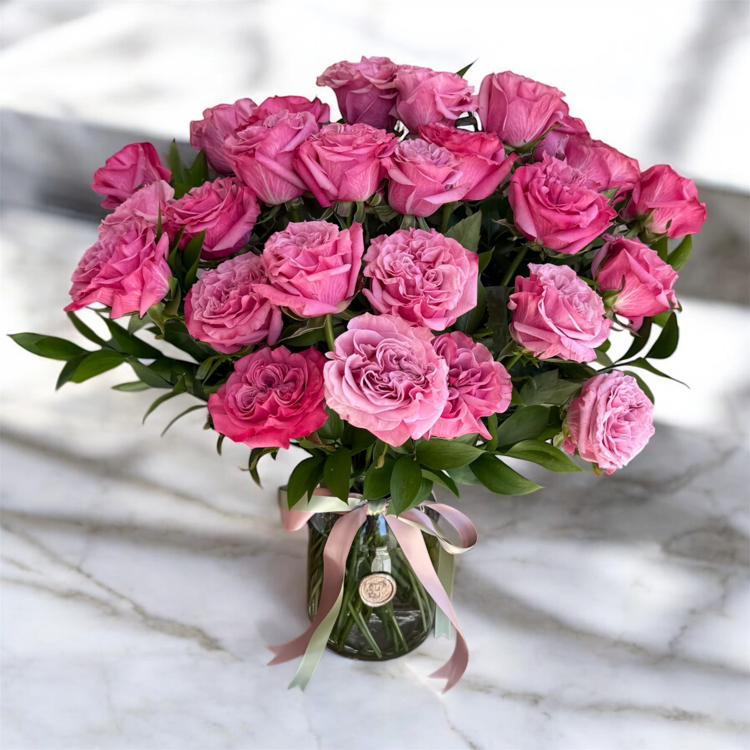 Blushing Hearts - Filled with charm and elegance, &quot;Blushing Hearts&quot;. features pink roses in soft, romantic hues and delicate ruffle textures. Accented by fresh greenery, it's arranged in a clear glass vase and finished with a ribbon and our signature wax seal. Perfect for celebrating love, friendship, or life's special moments. this arrangement adds beauty and meaning to any occasion.