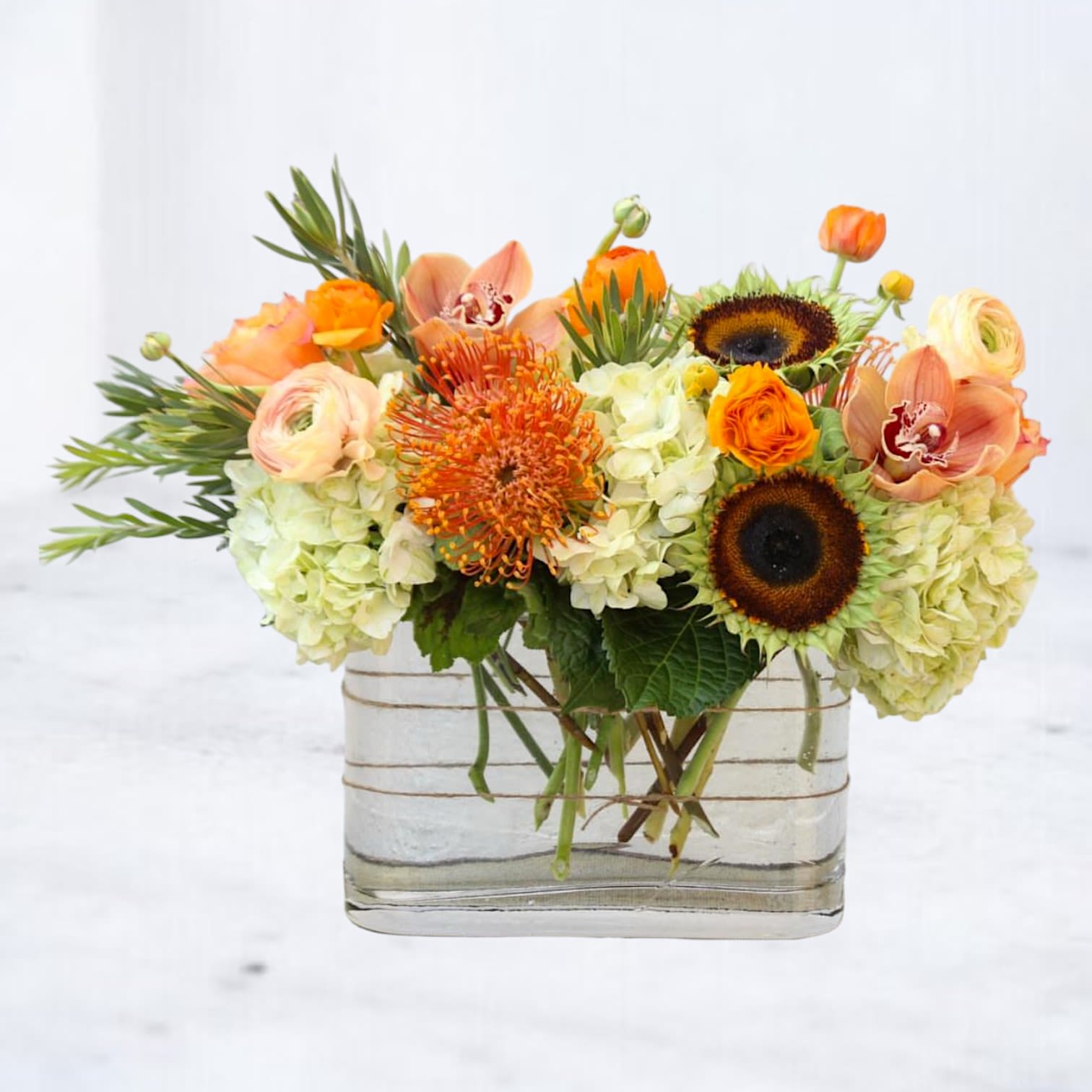 Burnt Citrus - Radiating heat, texture, and vibrancy, Burnt Citrus is a modern floral arrangement that blends exotic warmth with dynamic structure. Bold sunflowers, fiery pincushion protea, peachy ranunculus, and vivid cymbidium orchids are anchored by lush green hydrangea for a striking palette that feels both grounded and electric.  Designed in a contemporary rectangular vase, this statement piece is perfect for birthdays, housewarmings, or any moment that calls for unforgettable color and form. Available for same-day delivery in Houston and surrounding areas.
