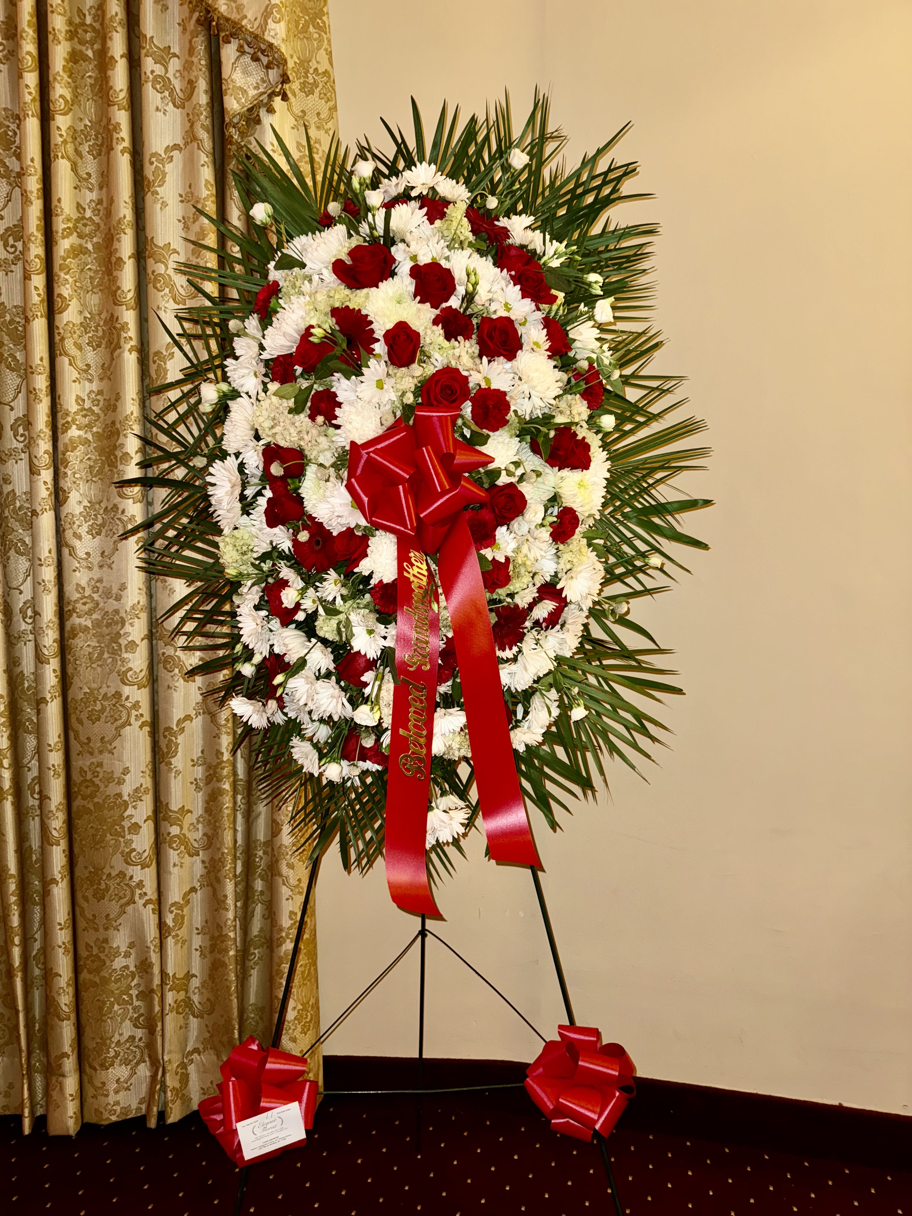 Standing spray red and white.  - A dignified tribute of love and remembrance, this standing spray blends the elegance of red roses with the warmth of red carnations and the purity of white blooms. Framed with lush greenery and accented by a bold red ribbon, it offers heartfelt comfort and honor in times of farewell.