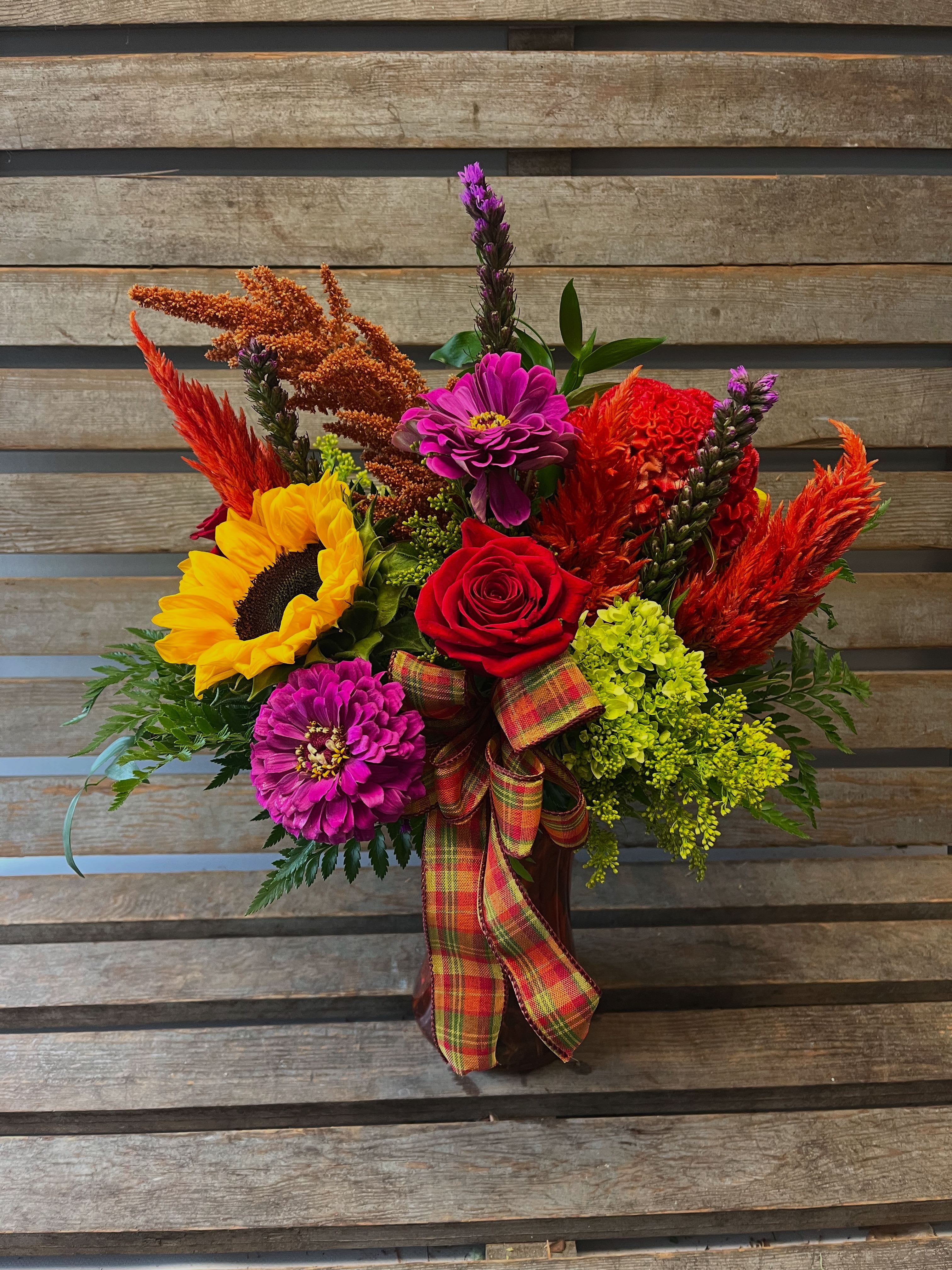 Perfectly plaid fall arrangement Perfectly plaid fall arrangement - A colored glass gathering vase filled with designer’s choice of local seasonal fall flowers. Vivid colors! Plaid bow added just for fun!