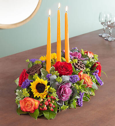 Garden Of Grandeur Centerpiece -  Autumn gatherings call for a grand centerpiece. We’ve designed our luxurious arrangement with a mix of brightly colored blooms and unique, textural accents, creating a truly unique table display. A trio of orange candles provides the perfect finishing touch for spirited celebrations.