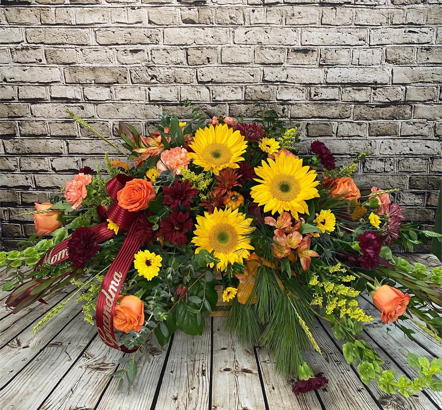 Autumn Memories Casket Spray - This vibrant casket spray offers a heartfelt expression of love and remembrance with the rich, warm tones of autumn. Golden sunflowers, deep burgundy mums, and bright yellow daisies are beautifully paired with orange roses and soft rust-toned blooms. Accented with fresh greenery and seasonal textures, this design creates a radiant display of color that symbolizes comfort, gratitude, and cherished memories. Perfect for honoring a loved one with beauty and warmth, this traditional spray brings both brightness and dignity to the service. Blooms may vary.   Approximate Dimensions: 30&quot;w x 12&quot;H 