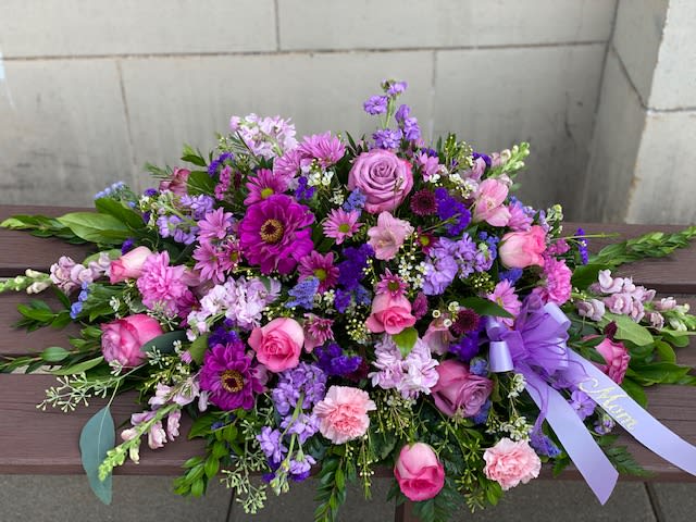 Compassionate Lavender Blooms Casket Spray - Honor her femininity and generous spirit with a blanket of sweetly scented lavender, pink and purple flowers that create a magnificent final tribute, sincerely expressing devotion and love. Gorgeous flowers such as lavender and pink roses, pink snapdragons and alstroemeria, purple and lavender chrysanthemums. All are entwined with eucalyptus, huckleberry and ming fern.