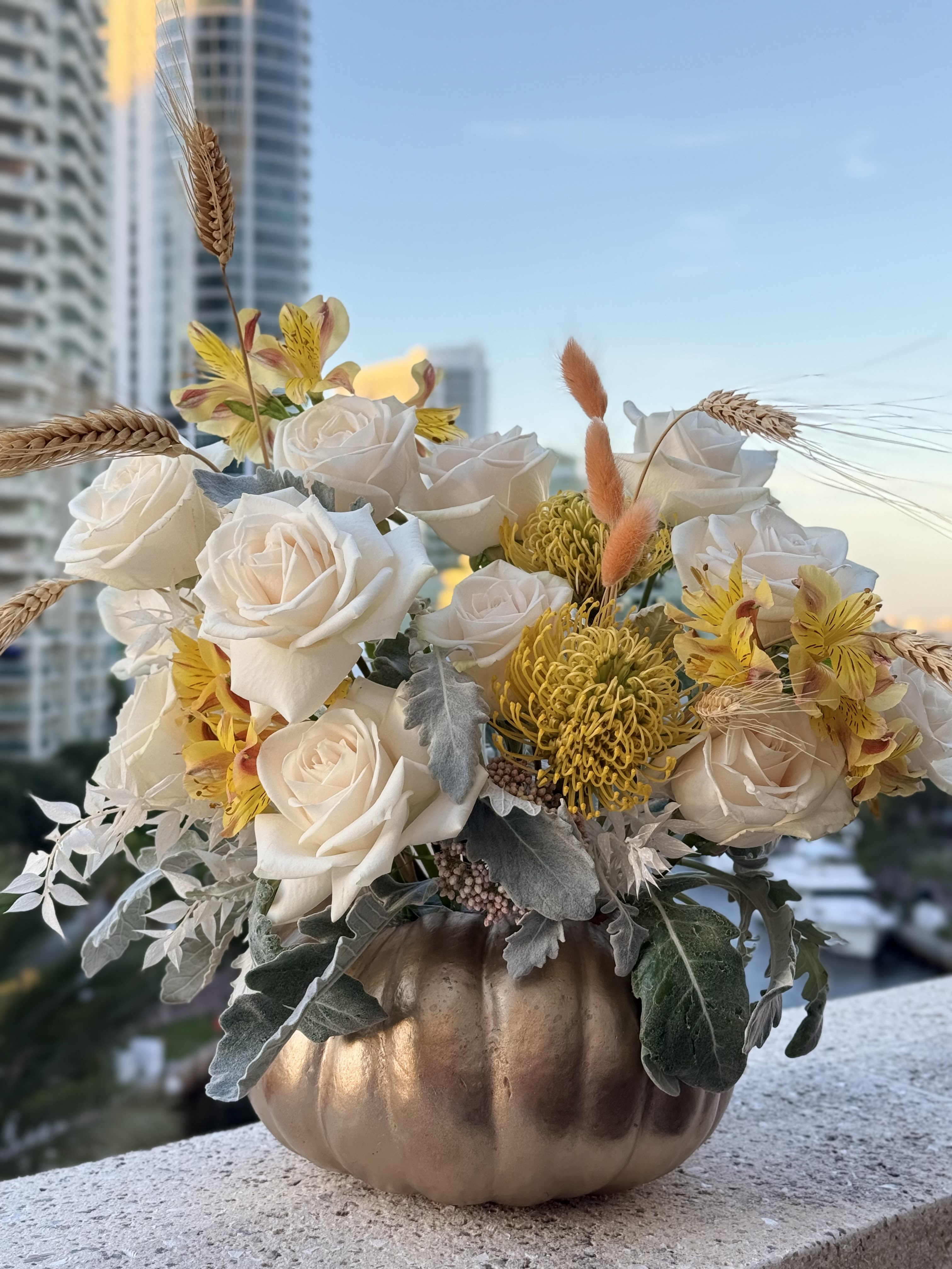 Autumn Glow - Creamy white roses and golden accents intertwine with natural wheat in a rustic pumpkin. A radiant arrangement that reflects the abundance of the harvest and the warmth of autumn gatherings. Flower Type: Creamy white roses, Yellow alstroemeria, Golden pincushion protea, Natural wheat stalks  Arrangement set in a natural pumpkin  