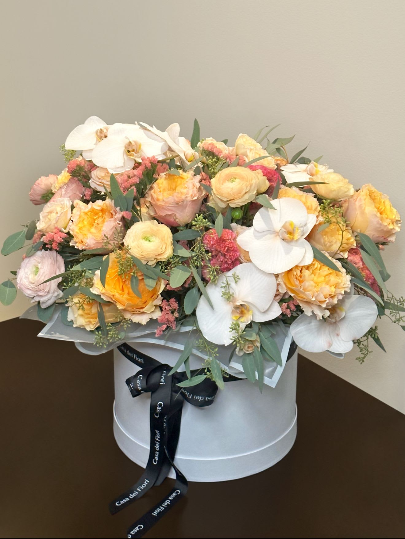 Soleil Bloom - A luxurious hatbox arrangement overflowing with golden garden roses, creamy orchids, and blush ranunculus, accented by soft greenery and pops of coral, evoking warmth, elegance, and celebration.