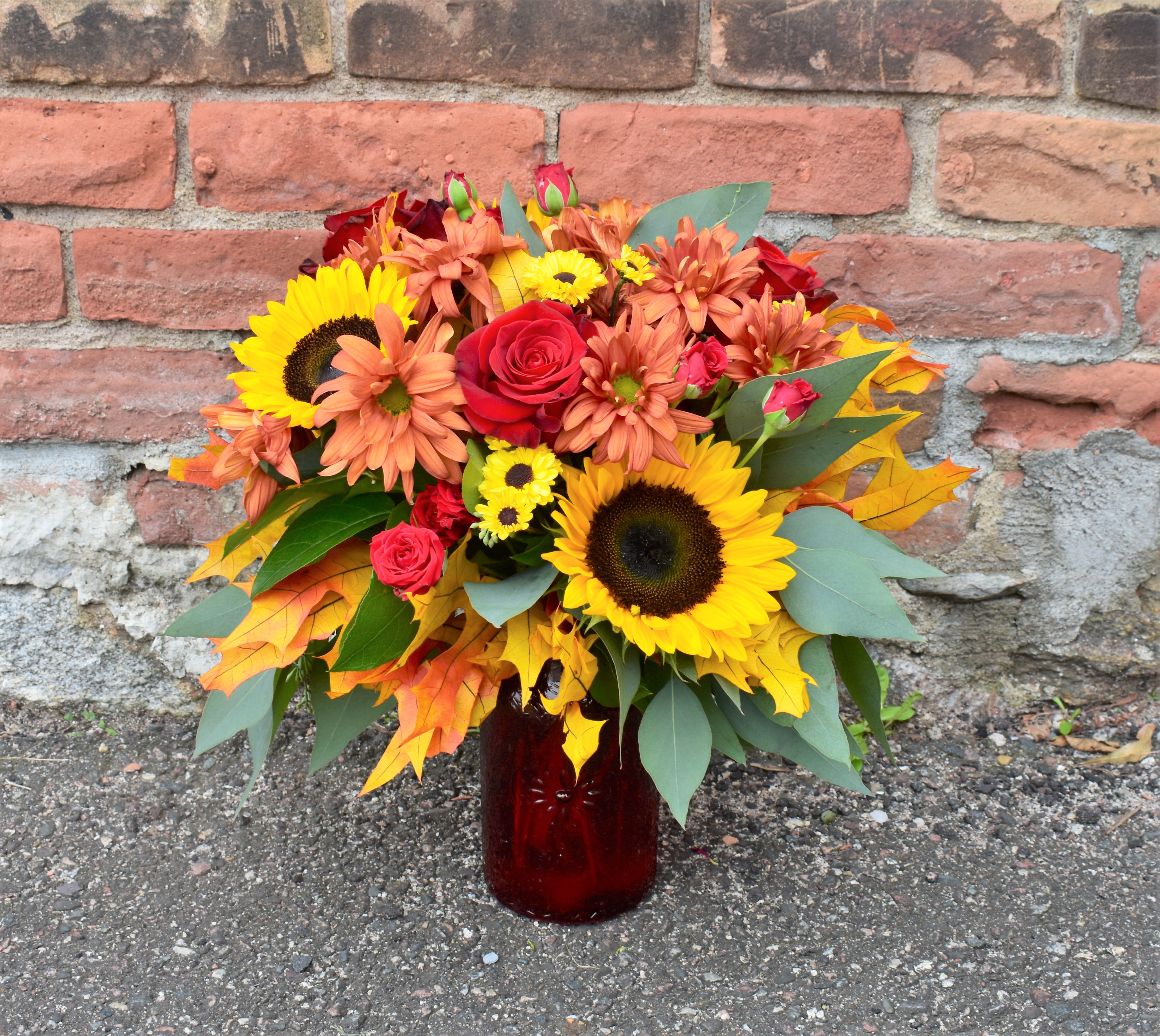A Walk through Leaves - Celebrating the vibrant colors and textures that fall brings, this bouquet is perfect for celebrating any occasion! Bright sunflowers, yellow and bronze mums, red roses, and eucalyptus is accented by fall leaves and designed in a crimson glass vase. Bring the beauty of an outdoor autumn day into your home! 