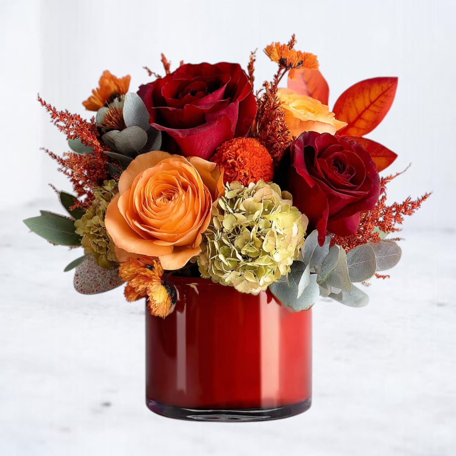 Fireside Radiance - Bring the glowing warmth of autumn into any space with our Fireside Radiance bouquet. This rich seasonal arrangement features deep crimson and vibrant orange roses, hydrangea, and fiery bronze accents, all artfully arranged in a sleek 4x4 red vase. The bold hues and lush textures evoke the comfort of sitting beside a crackling fire on a crisp fall evening.  Perfect for fall gatherings, holiday décor, or as a heartfelt gift, Fireside Radiance is a stunning reminder of the season’s beauty and warmth.