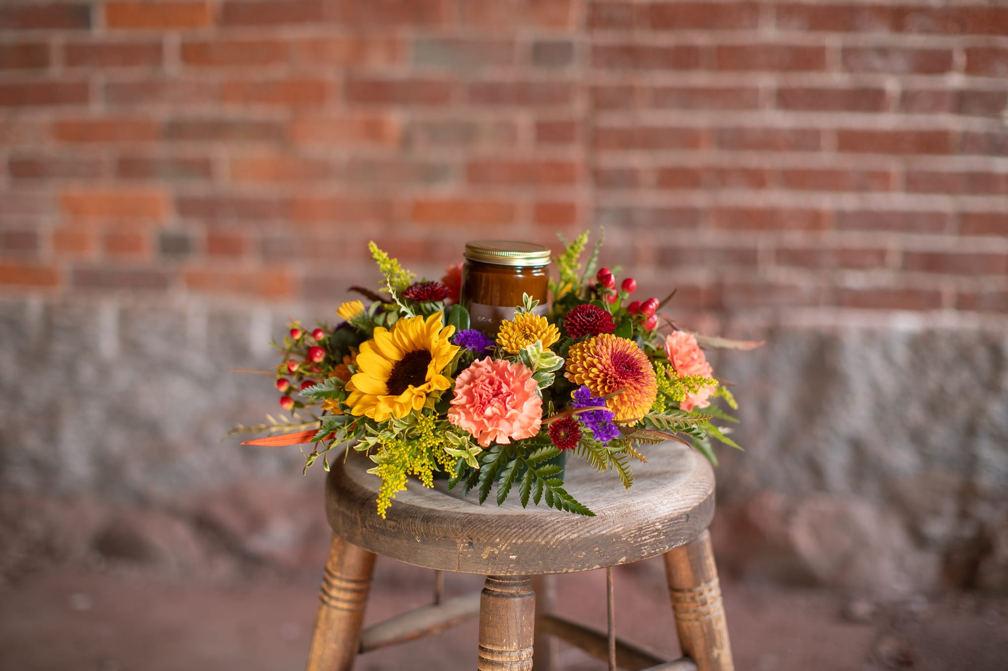 Season Scented Centerpiece - Illuminate the senses of Thanksgiving spirit with the delicate fragrant candle centerpiece. The amber jarred candle is handcrafted. The scent of seasonal apple and pumpkin is perfect for harvest festivities. The glowing candle is surrounded by beautiful rich autumnal toned flowers. Sunflowers, roses, mums, poms and carnations. Seasonal foliage of seeded eucalyptus and grevilia complete the look to accompany joyous fall gatherings.