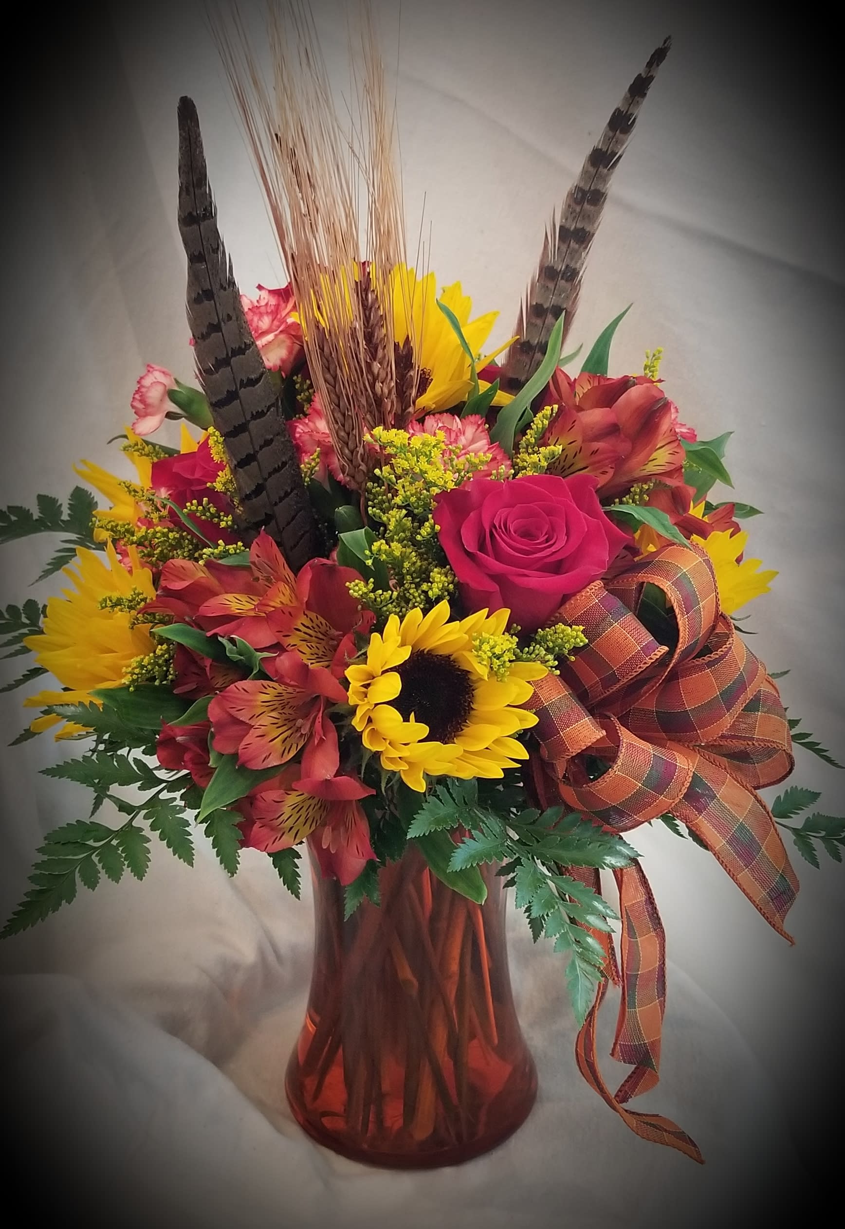 Harvest Vase - A traditional arrangement of roses, sunflowers asters, alstroemeria and other flowers accented with dried wheat and pheasant feathers in a clear vase.  