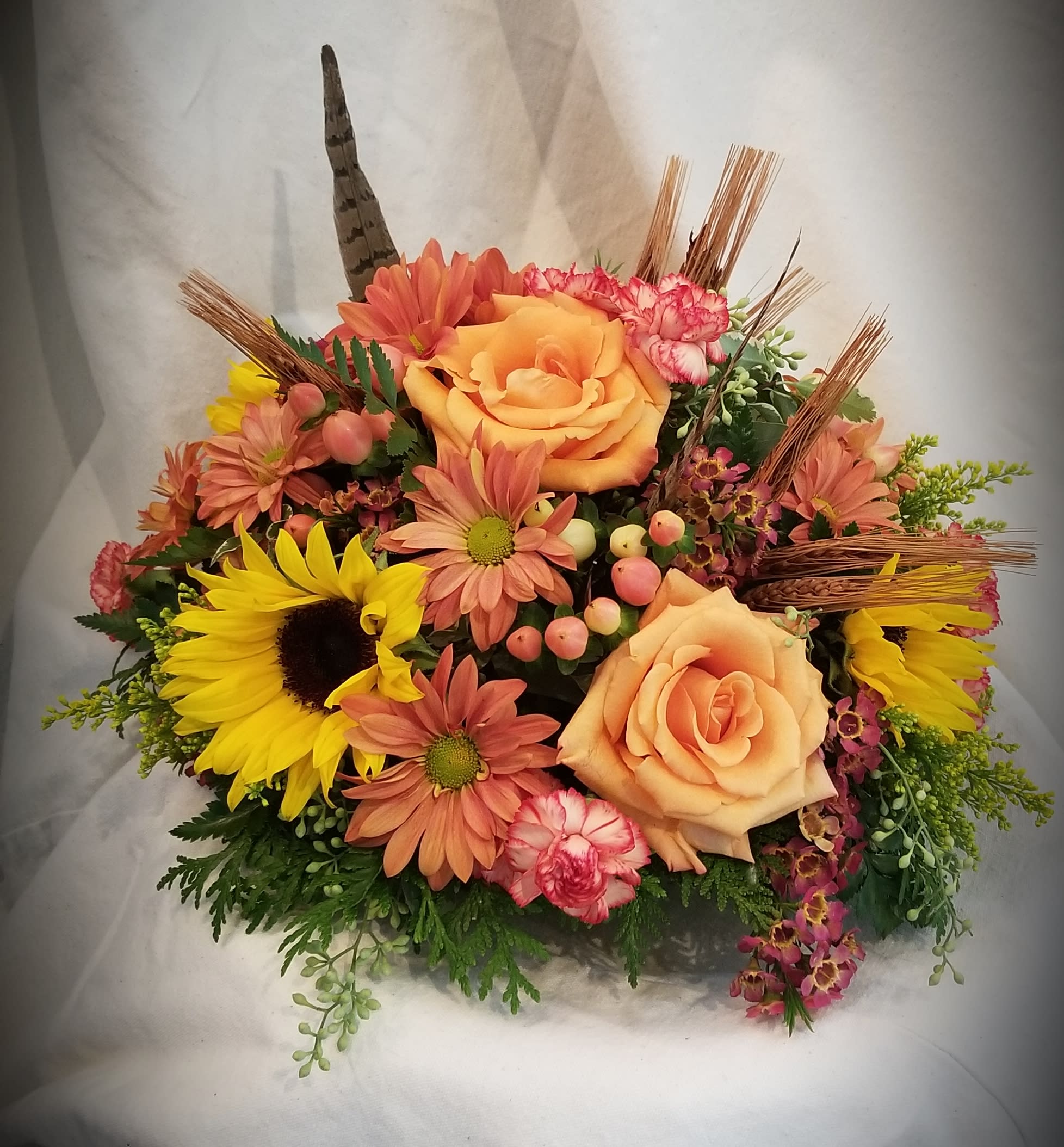 Autumn Centerpiece - Arranged in dish in autumn colored flowers, this round centerpiece measures about 9&quot; in height by about 12&quot; in diameter.  It includes orange roses, hypericum, daisies, sunflowers, mini carnations, dried wheat and pheasant feathers.
