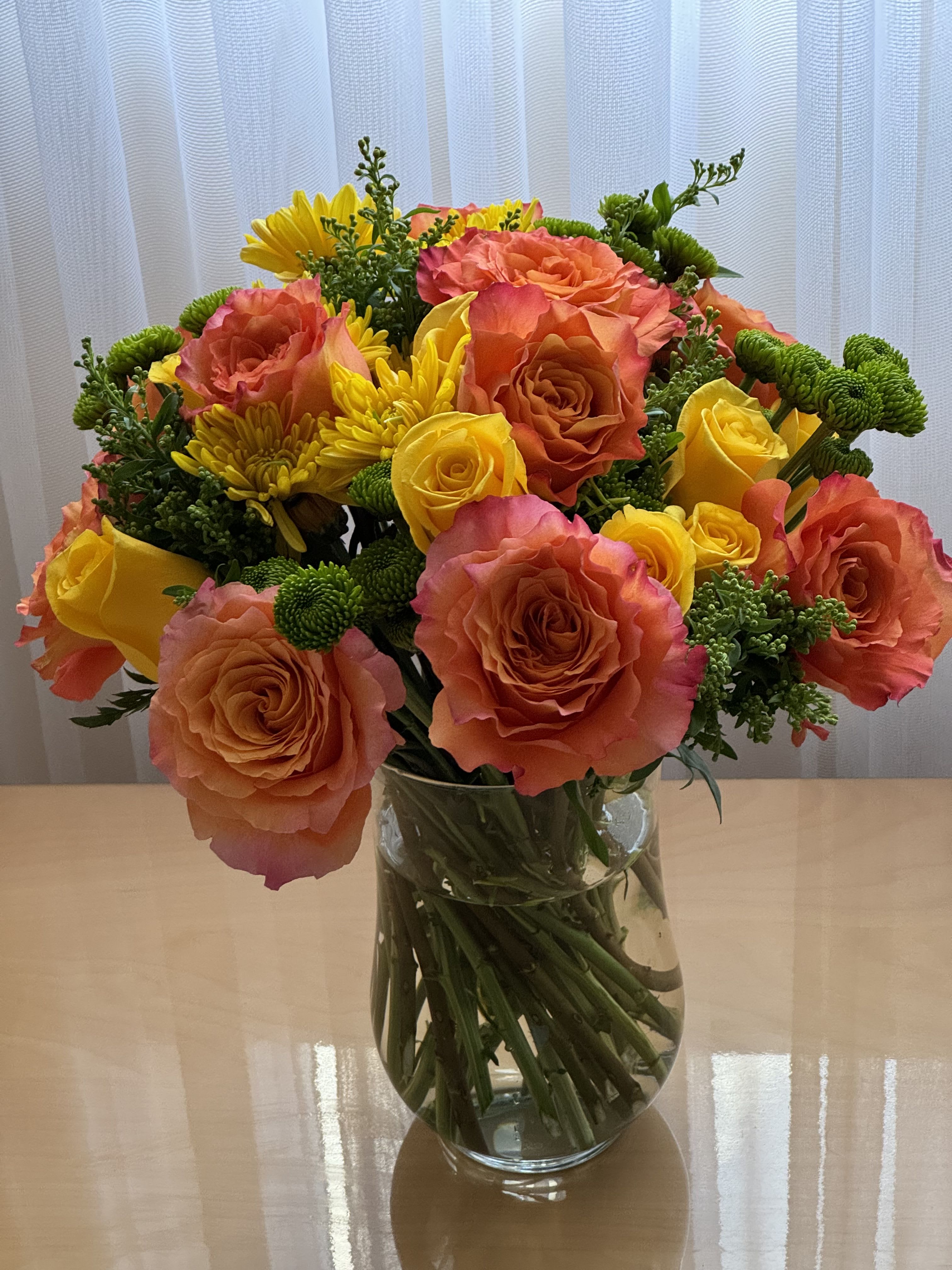 Joyful Bloom Bouquet - This vibrant and cheerful floral arrangement is perfect for brightening up any space or occasion. It features a delightful mix of orange and yellow roses, complemented by sunny yellow chrysanthemums and lively green button poms. The flowers are artfully arranged in a clear glass vase, allowing the natural beauty of the stems to add a touch of elegance. The combination of warm, vivid colors and fresh greenery creates a harmonious and uplifting display that is sure to bring joy to anyone who receives it. It is ideal for birthdays, anniversaries, or simply as a thoughtful gesture to show you care.  Orange Roses Yellow Roses Yellow Mums Greens  Height 14 inches Width 8 inches