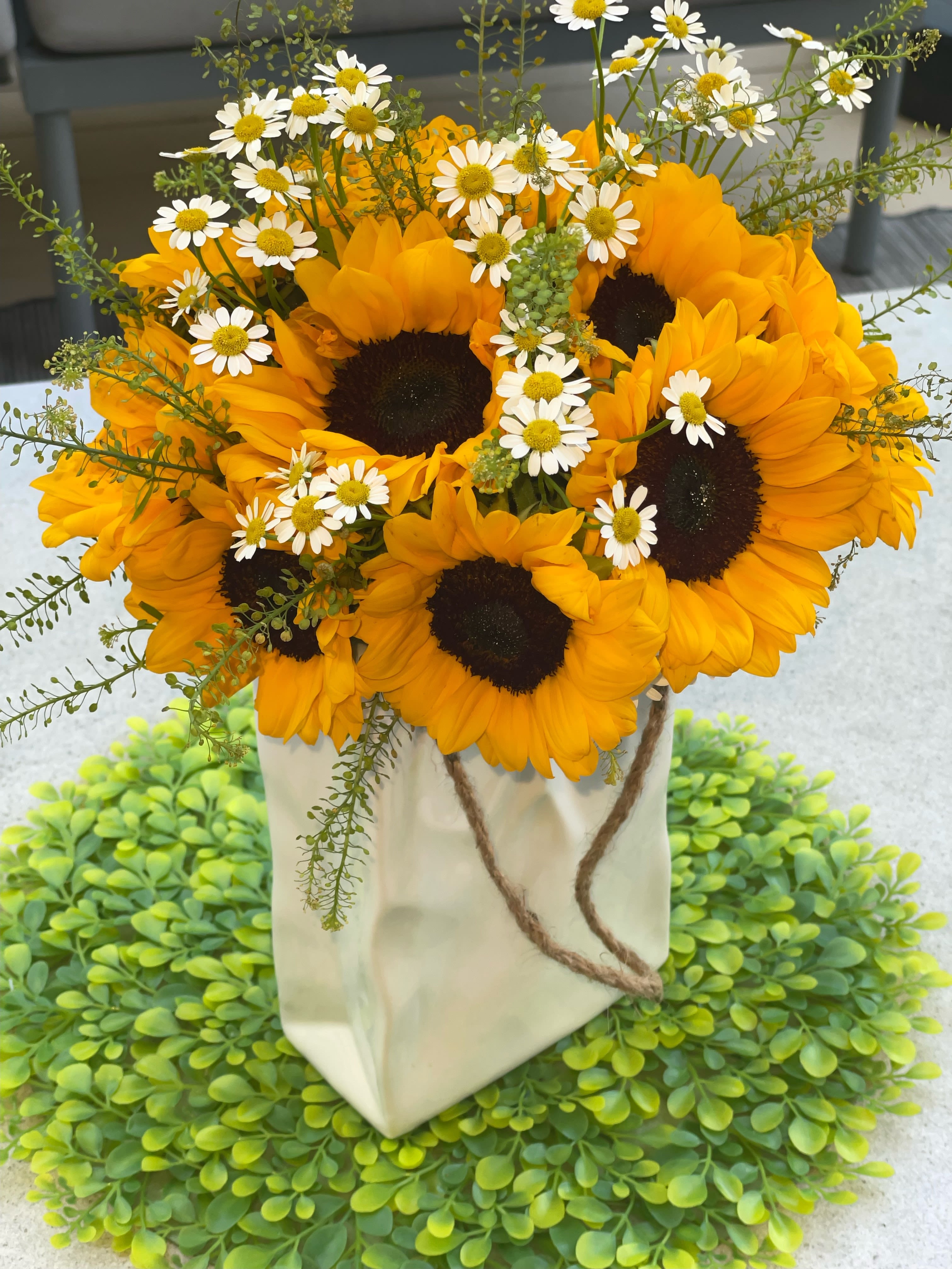 Sunshine Delight Bouquet - This stunning floral arrangement features a beautiful blend of sunflowers and daisies. The vibrant yellow sunflowers, with their large, dark centers, create a striking contrast against the delicate white daisies scattered throughout. The arrangement is enhanced with sprigs of greenery, adding a touch of natural elegance. The flowers are artistically arranged in a chic, eco-friendly white paper bag with rustic rope handles, making it a perfect gift or centerpiece for any occasion.  Sun Flowers Daisies  Height 15 inch Width 15 inch