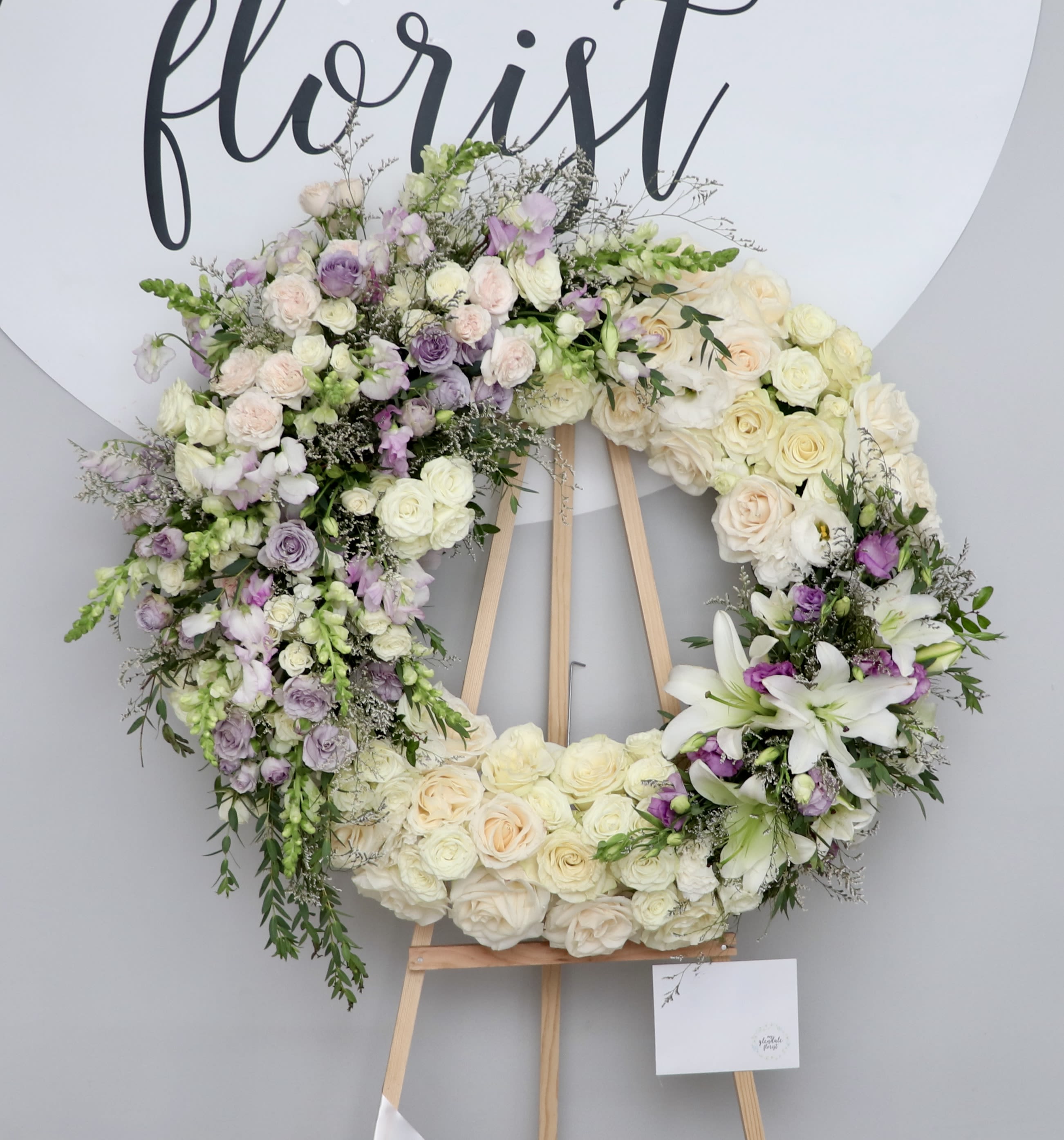 Lavender Harmony Wreath - This beautiful sympathy wreath is made with white roses and accented with two adjacent lavender designs. Snaps, lilies, roses, and other seasonal blooms are included to create harmony and balance. Each upgrade adds more flowers!  Standard size is 30'', deluxe is 36'', and premium is 42''.  We include easel, printed banner and delivery (some fees may apply). An additional banner can be added for $15.