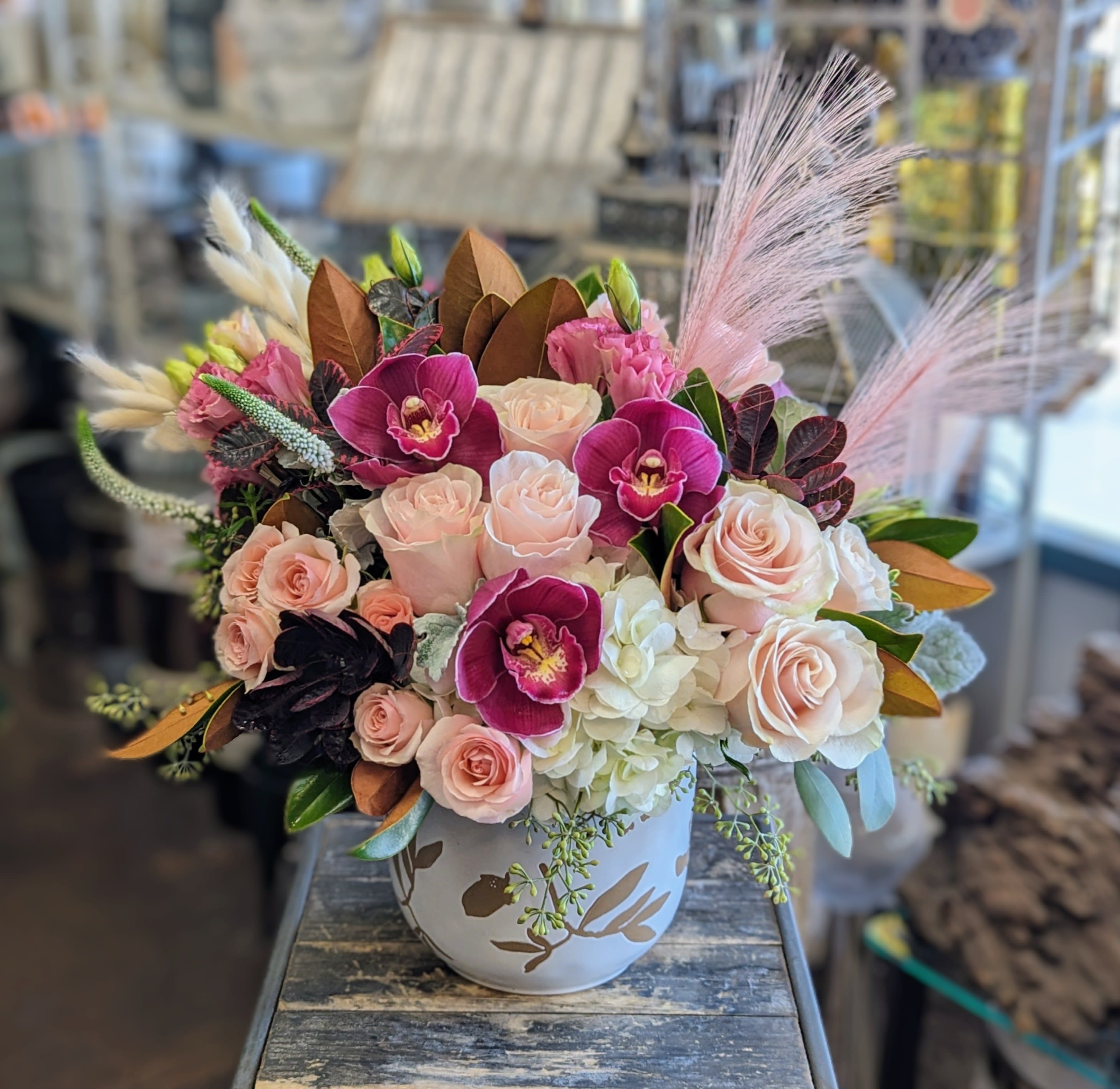 Clove Clove - A mix in shades of pinks and burgundy, accented with magnolia foliage, arranged in an autumn themed vessel.