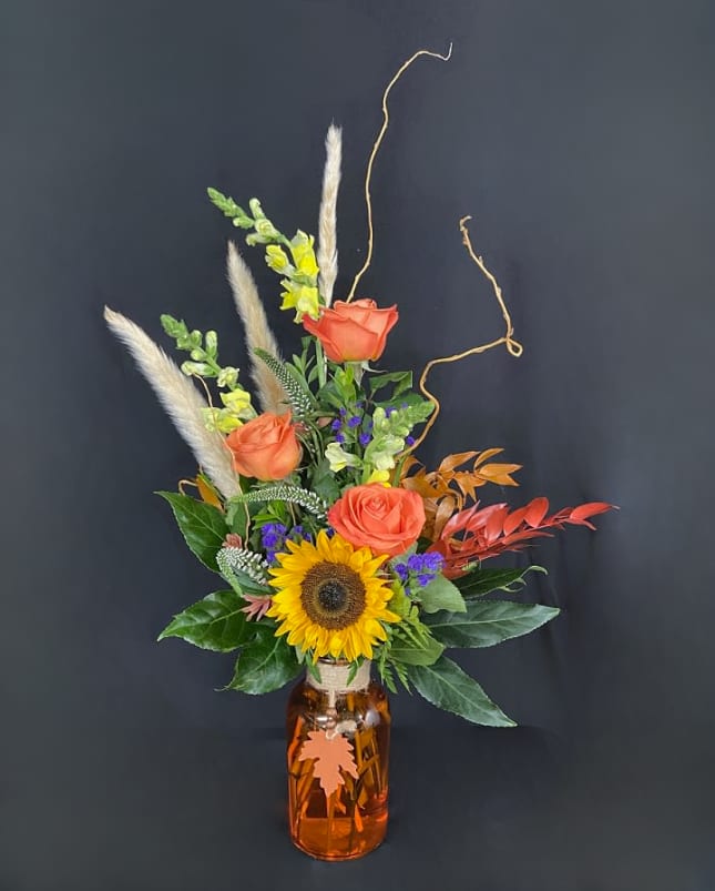 Autumn Leaves - It feels like Autumn once the first leaves fall! Our Autumn Leaves arrangement brings this to life with an assortment of fall colored roses, sunflowers, veronica, snapdragons, statice and daisies accented with preserved Italian ruscus leaves, aralia leaves, plumas grass, and curly willow branches in a burnt orange glass vase with decorative oak leaf medallion. 