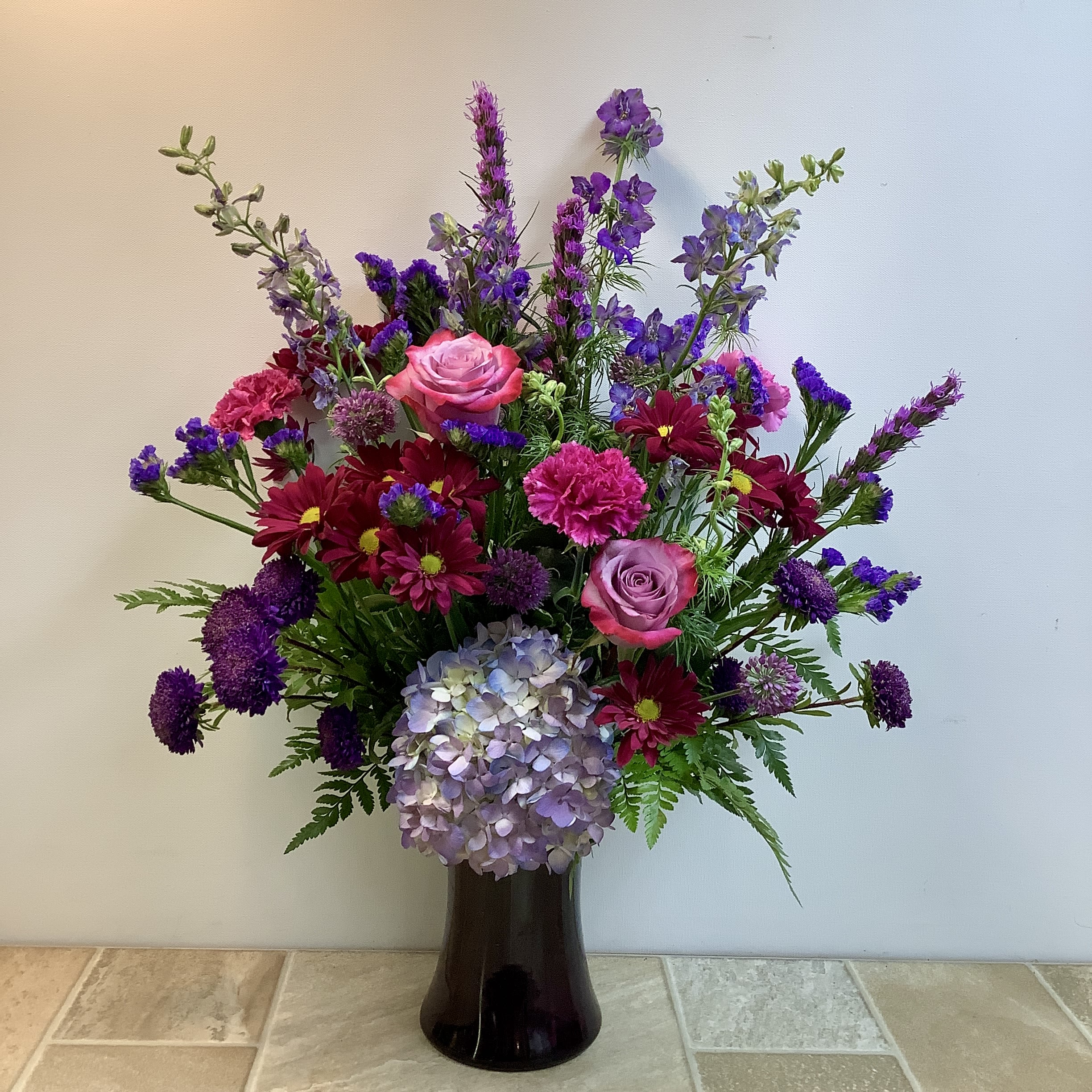 Seth Seth - This large, abundant floral arrangement features a vibrant mix of pink and purple flowers with splashes of green foliage. A lush hydrangea takes center-stage, surrounded by purple daisy mums and statice, lavender roses, and pink carnations. Accent blossoms include liatris, larkspur and more. Approximate size: 20" w x 27" h Primary flowers: hydrangea, mum, rose, carnation