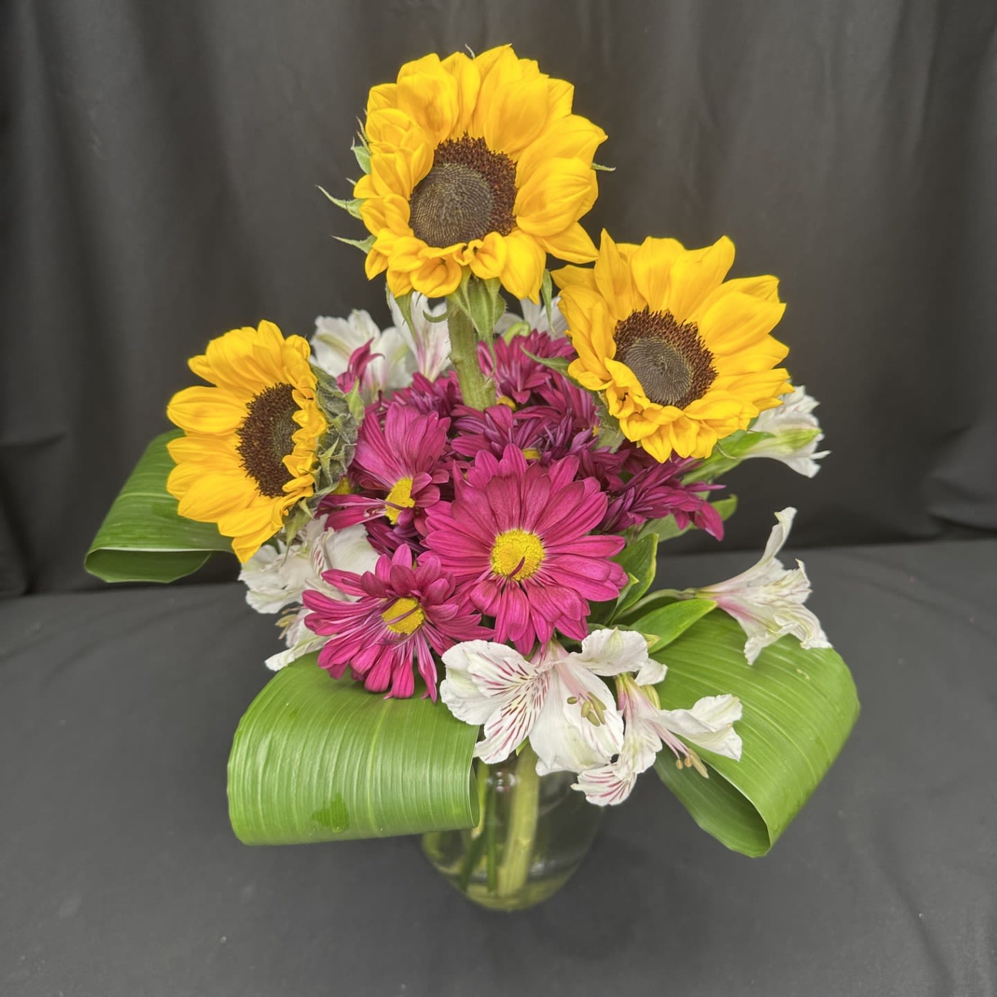 Golden Harvest - Bright and joyful, this arrangement pairs radiant golden sunflowers with bold fuchsia daisies and soft white alstroemeria, all framed by glossy green leaves. Bursting with color and texture, it brings the warmth of a golden harvest field into any space. Designed in a clear vase, it’s a cheerful centerpiece perfect for celebrating the beauty of the season.