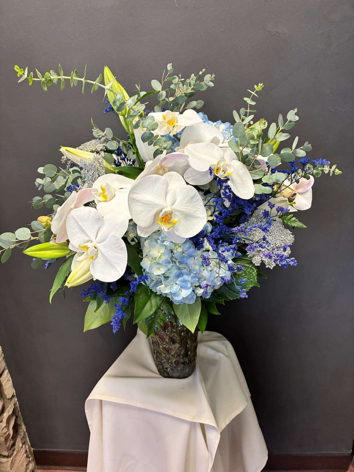 Whispers of Elegance - A serene symphony of soft hues and graceful textures, this arrangement blends classic refinement with a touch of whimsy. Cascading white orchids anchor the design with sculptural beauty, lilies and light blue hydrangeas add gentle warmth and cool contrast. Delicate filler blooms in blue and white dance among eucalyptus sprigs and lush greenery, creating a layered, airy composition. Each bloom radiates with quiet sophistication—ideal for weddings, upscale events, or heartfelt gifts that speak volumes without saying a word.