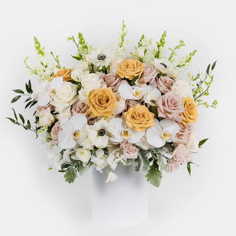 Creme de la Creme  - This stunning floral arrangement showcases a romantic mix of gold, blush and creamy white roses, white anemones, accented with white orchids and dusty miller for added elegance. Featured in White Floral Box 8&quot;.