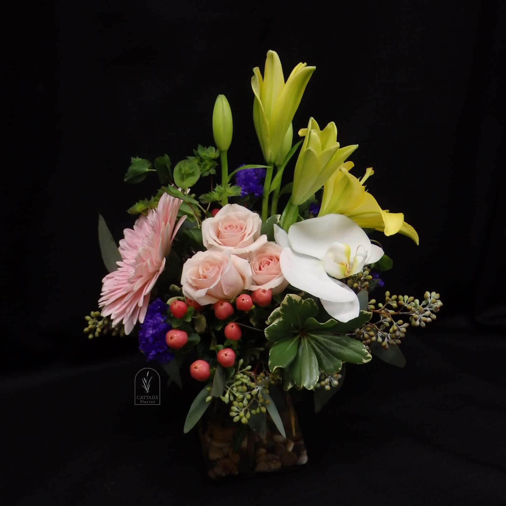 Tiny Dancer - “Tiny Dancer” is a sweet mix of soft spray roses, cheerful yellow lilies, a single elegant white orchid, and playful accents of gerbera daisies, berries, and greenery—arranged in a compact vase for a charming and uplifting display. 