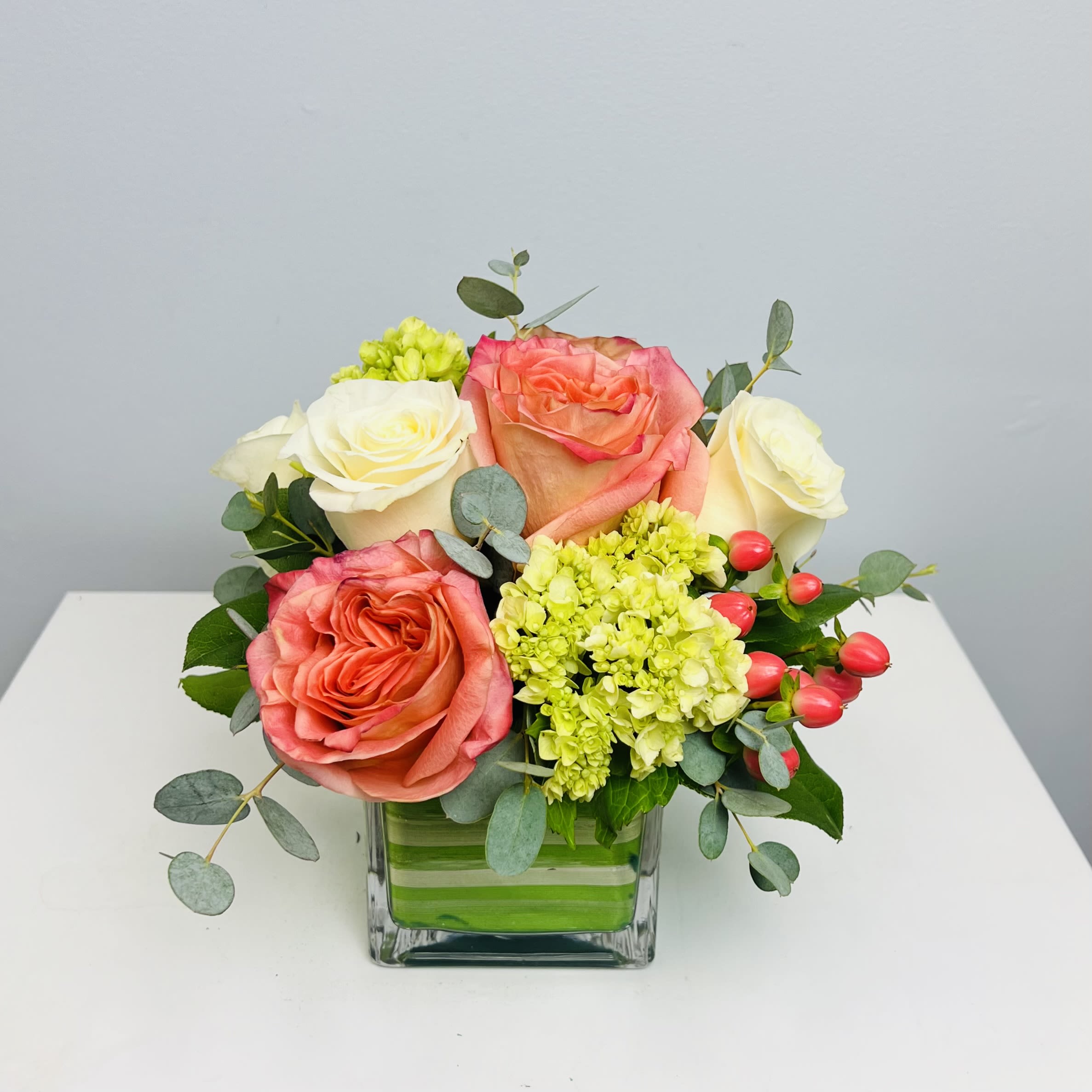 Grace - Simple elegance and refinement defines Grace. Beautiful soft tone roses are accented with green miniature hydrangea, and pink berries in a clear leaf wrapped cube vase.   Approximately 10” Tall 