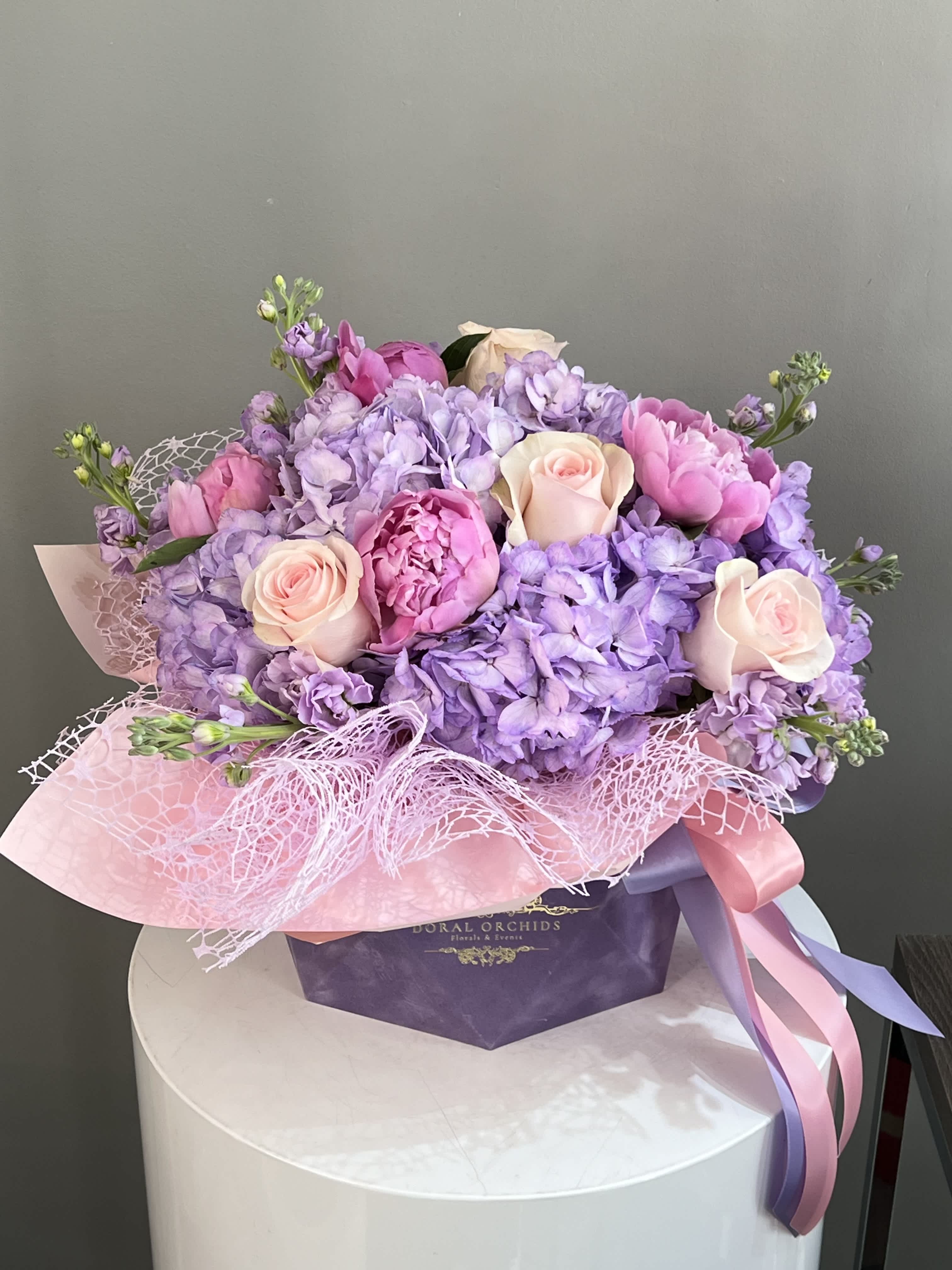 Enchanted Gardens ™ Enchanted Gardens ™ - This dreamy arrangement combines lavender hydrangeas, soft blush roses, and rare pink peonies, creating a luxurious pastel palette. Wrapped in delicate lace and satin ribbons, it’s a stunning statement of grace and elegance.