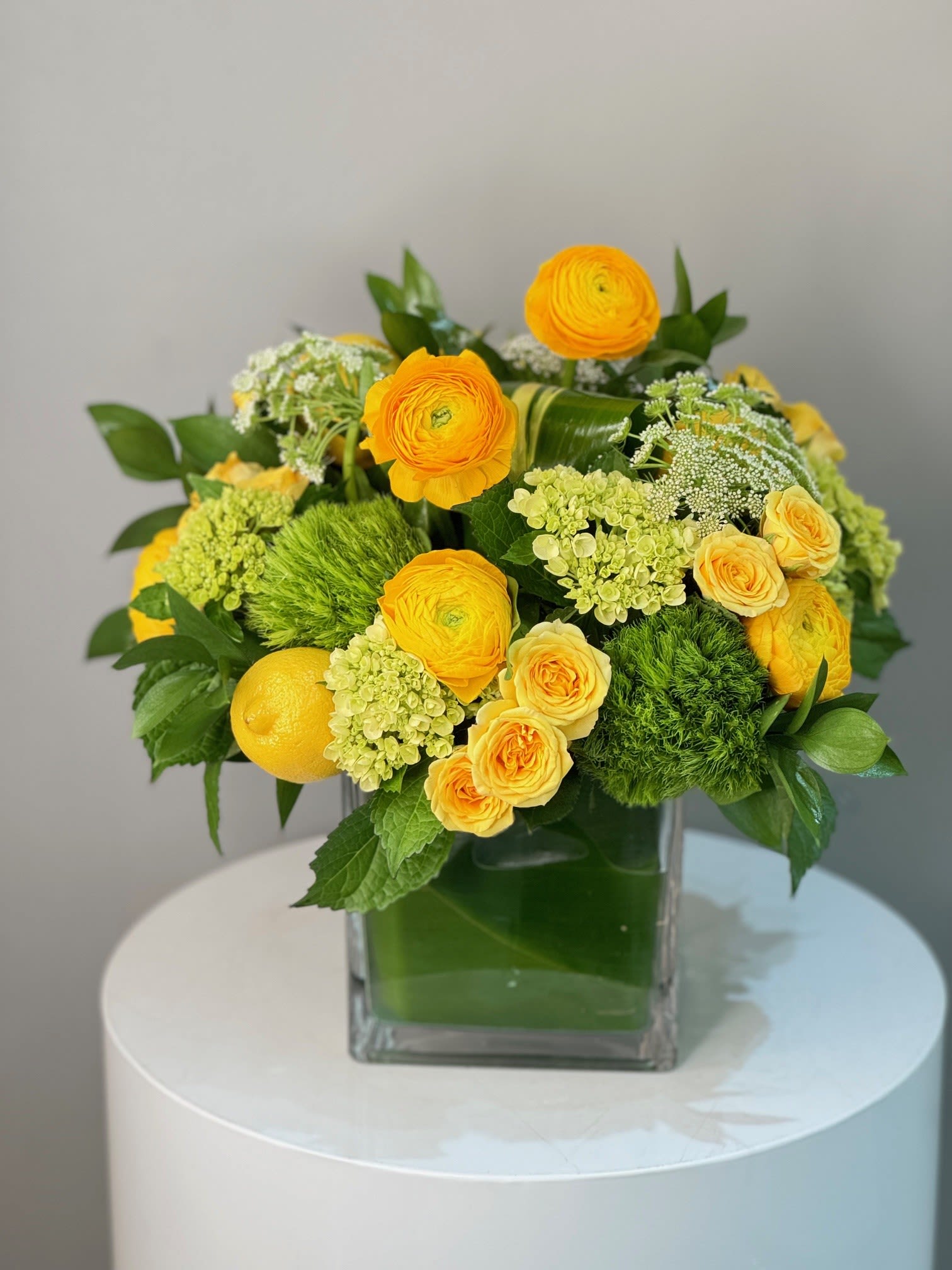 Lemon Twist ™ - This arrangement bursts with sunny yellow ranunculus and roses, paired with fresh green hydrangeas and lush foliage for a lively, cheerful display. Presented in a modern glass vase, it radiates brightness and warmth—perfect for bringing joy to any space.
