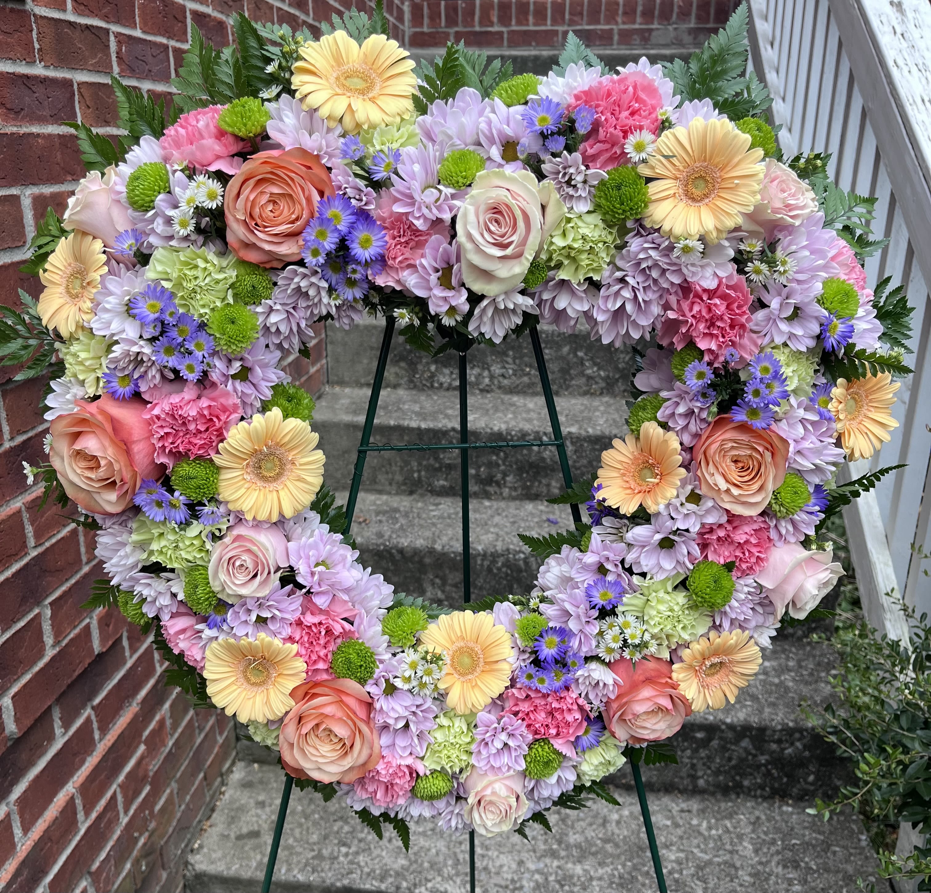 Eternal Day - This beautiful heart easel has pink and green carnations, pink and peach roses, peach Gerber daisies, purple daisies, green buttons, purple and white Monti and baby's breath with greens.