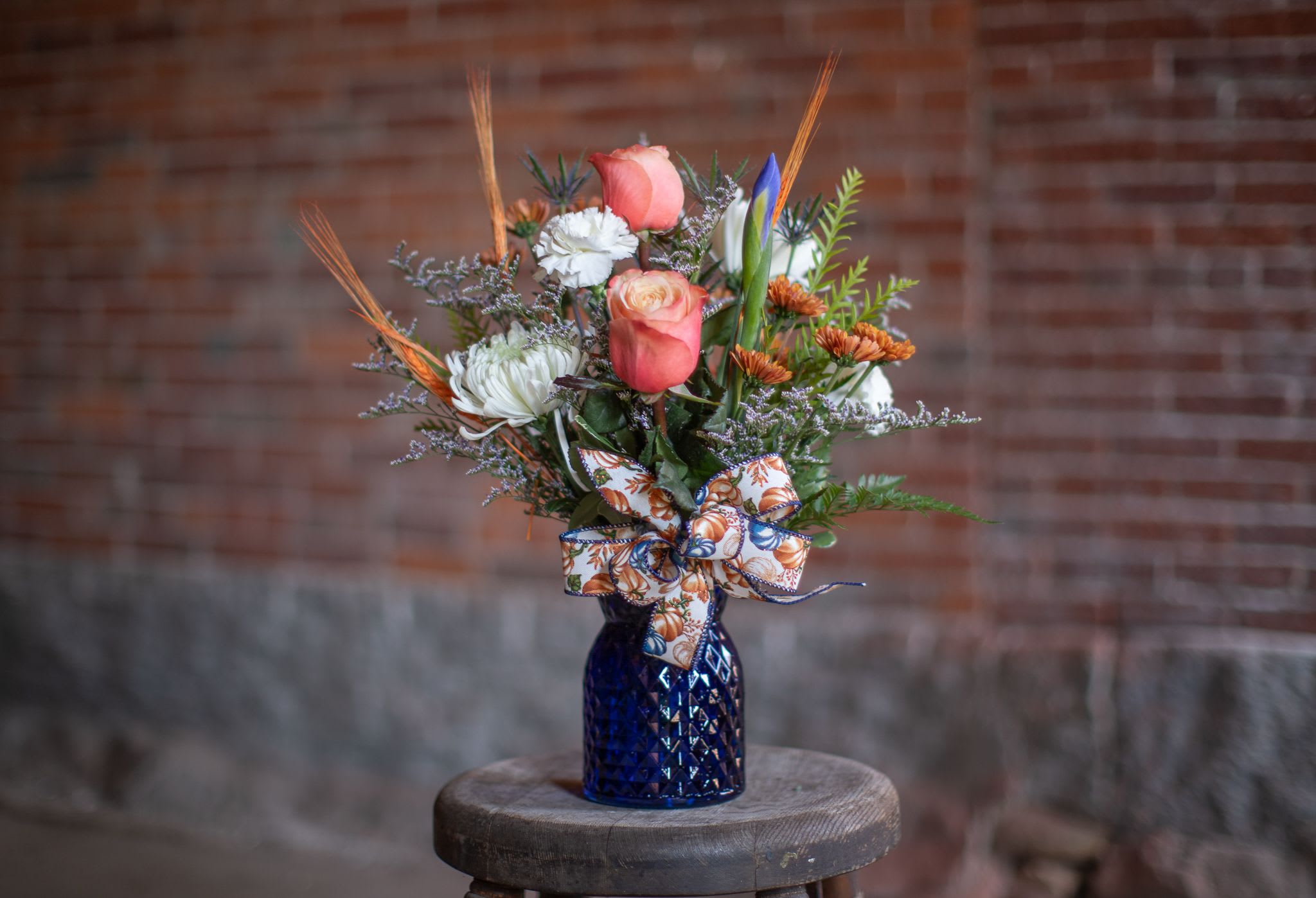 Country Pumpkins - Festive fall favorites with a blue twist. A cobalt blue vase is filled with designer blooms of cinnamon spiced blooms of iris, thistle, pom pons, cremons, roses or spray roses and more. A whimsical play on country bumkins is our latest fall design. Textural elements of wheat and thistle give this design all the fall-feels. The cinch vase design is completed with a handcrafted burlap bow of indigo and terracotta colored pumpkins. Flowers varieties may vary slightly from photo but over all color schematic will be created. Substitutions: In some instances we may need to use spray roses in place of the standard roses. We may use copper mums in place of white. Based on our inventory we may use blue poms in the place of irises.   