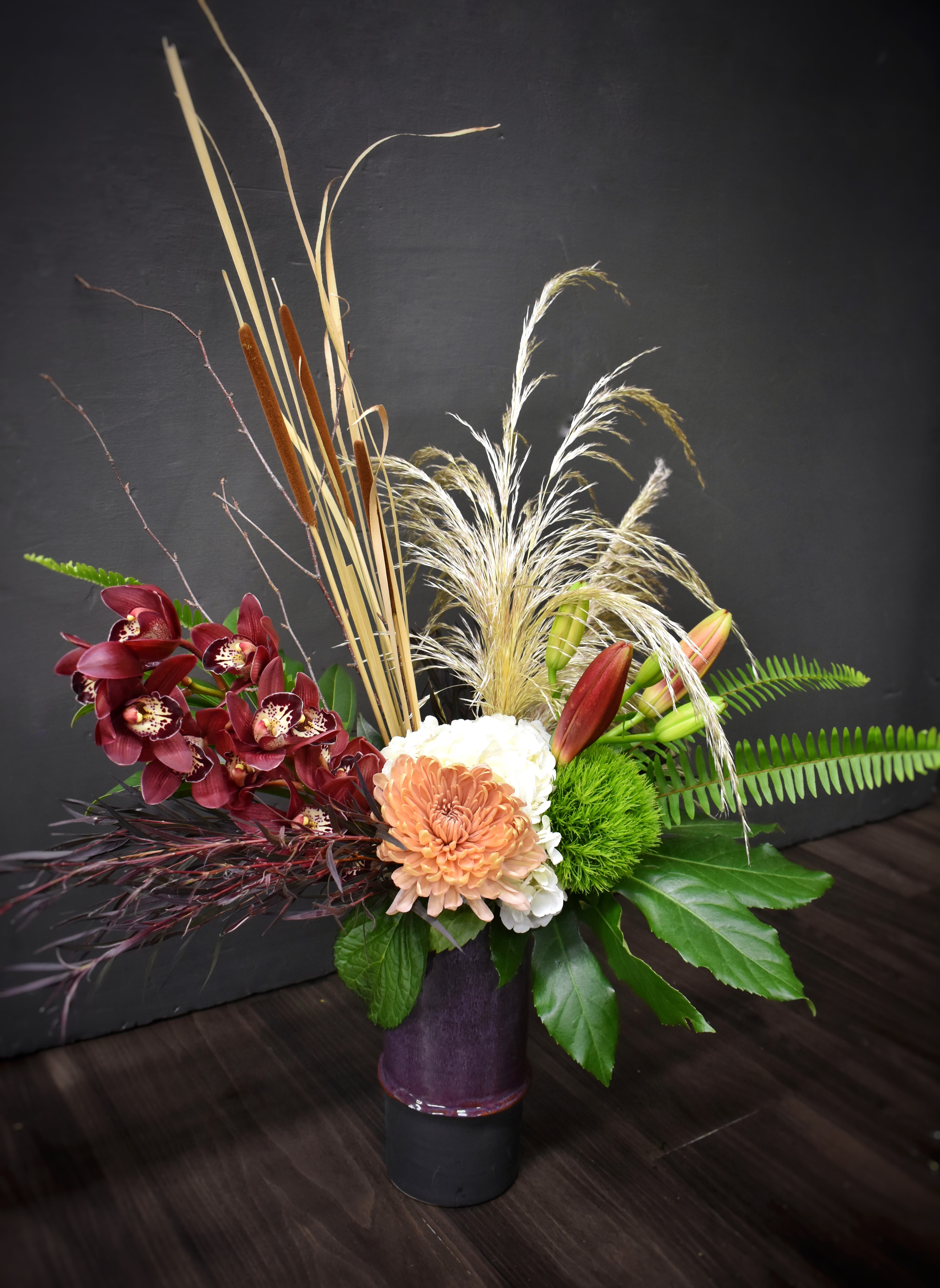 Orchid in Autumn - A little different than your average fall bouquet! This ceramic purple vase is exploding with seasonal pampas grass, cattails, rich green foliages, hydrangea, mums and a beautiful mini cymbidium orchid stem. Colors may vary.