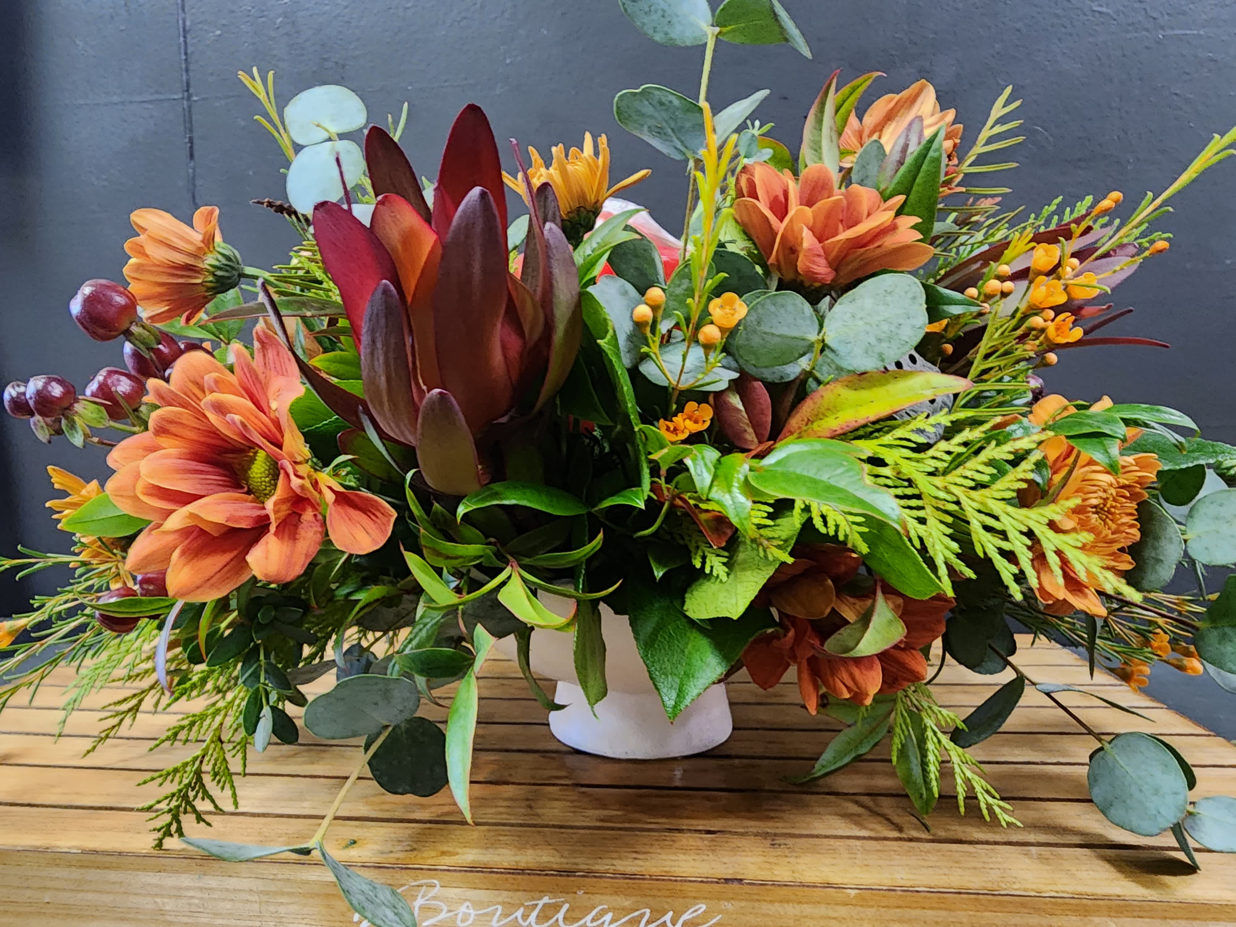 Thanksgiving Elegance with Pillar Candle - An all around flower arrangement designed with fresh flowers to include mix of fall chrysanthemums, daisies, mixes greens and leucadendron with a piller candle and in a pedestal container.  * Some stages of flower life may vary. (Example: Lilies may be more open than imaged.)  Flower shortages and product availability means that some substitutions will apply. Value will always be filled. This bouquet might include eucalyptus amongst the greenery. It may be toxic to pets including but not limited to Cats and Dogs if digested.