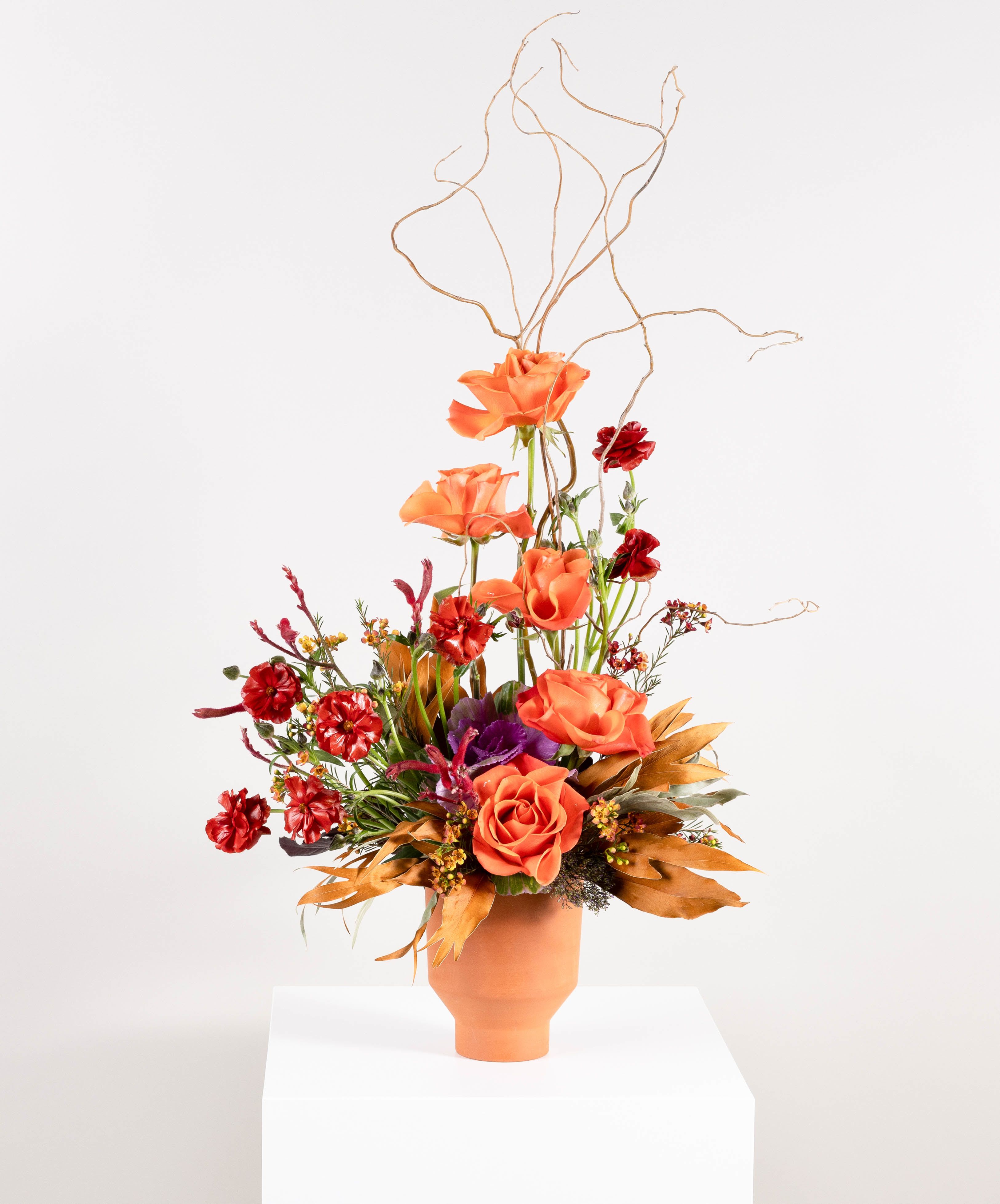 Good Thing Going - This elegant floral arrangement captures a dynamic balance of form and color, evoking a sense of graceful movement. Soft orange roses tower gracefully above a mix of deep red carnations, purple anemones, and rich green foliage. The rich warmth of the blooms is complemented by dried autumn leaves, which ground the composition with natural texture and depth. Above, sculptural twigs extend upward, creating an airy, upward flow that adds a touch of drama and elegance. The arrangement is presented in a simple terracotta vase, allowing the natural beauty of the flowers to take center stage.  Perfect for adding a touch of bold, yet refined beauty to any space, or as a thoughtful gift for someone who appreciates a blend of vibrant colors and organic, sculptural forms.