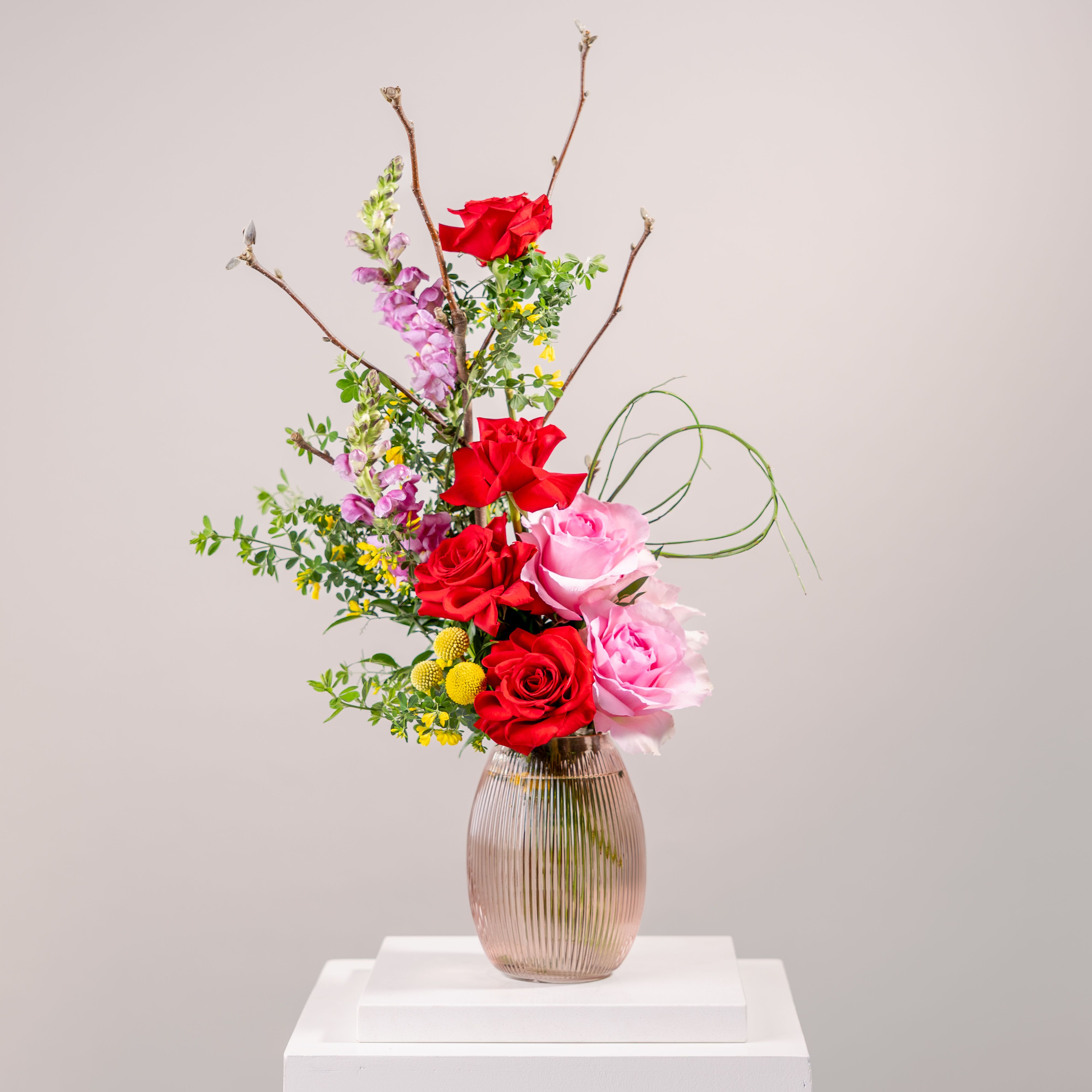 My Miss Mary  - This stunning floral arrangement showcases an artful blend of graceful forms and soft, refined hues. Lush red and pink roses, delicate pink snapdragons, and vibrant yellow blooms add a romantic and expressive touch, while thin woody branches extend upward, bringing movement and a sense of lightness. Presented in a classic ribbed pink glass vase, this arrangement radiates both modern charm and timeless sophistication—like a still moment captured in a blooming dreamscape. Perfect for adding a touch of contemporary elegance to any setting, or as a thoughtful gift for those who appreciate unique floral artistry. 