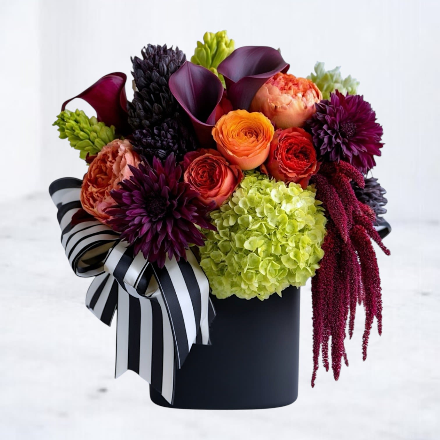 Haunted Harvest Bloom - The Haunted Harvest Bloom is a spellbinding Halloween arrangement designed to turn heads. Featuring fiery orange garden roses, lush lime green hydrangea, deep burgundy blooms, and sleek dark calla lilies, this bouquet captures the mystery and magic of the season. Accented with cascading amaranthus, bold textures, and a striking black-and-white striped ribbon, it makes a perfect centerpiece for Halloween parties, October birthdays, or simply celebrating fall in bold style. Arranged in a modern black vase, this luxe bouquet is available for Halloween flower delivery starting October 1st. 