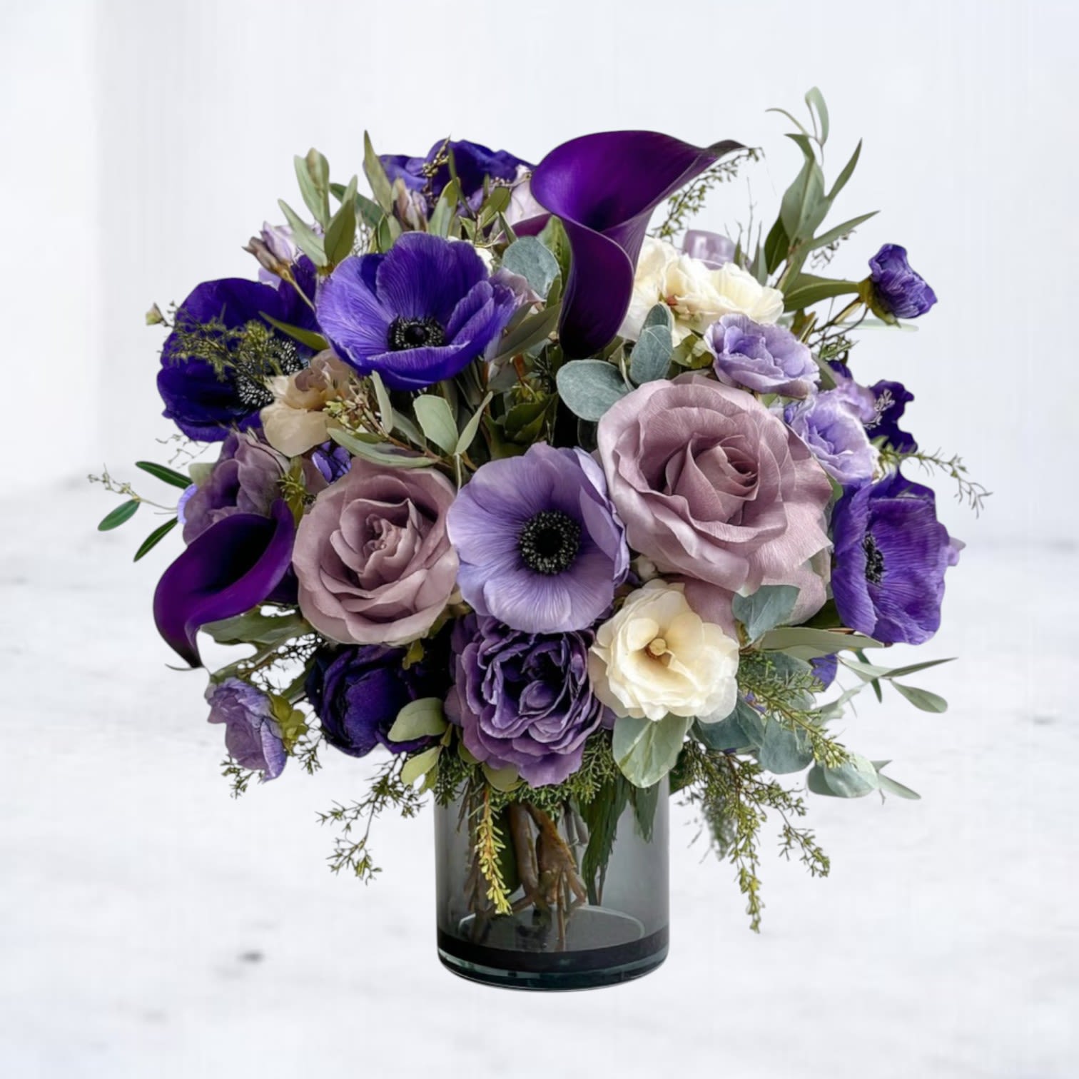 Enchanted Amethyst Bloom - The Enchanted Amethyst Bloom captures the allure of twilight with its captivating mix of deep purple calla lilies, soft lavender roses, and striking violet anemones. Accented with ivory blossoms and lush greenery, this modern arrangement is presented in a sleek glass vase for a sophisticated finish. Perfect for anniversaries, birthdays, or sending a touch of luxury just because, this bouquet brings an air of mystery and elegance to any setting. Available for same-day flower delivery in Houston and surrounding areas, this violet and lavender flower arrangement is a stunning choice for expressing love, admiration, or celebration.
