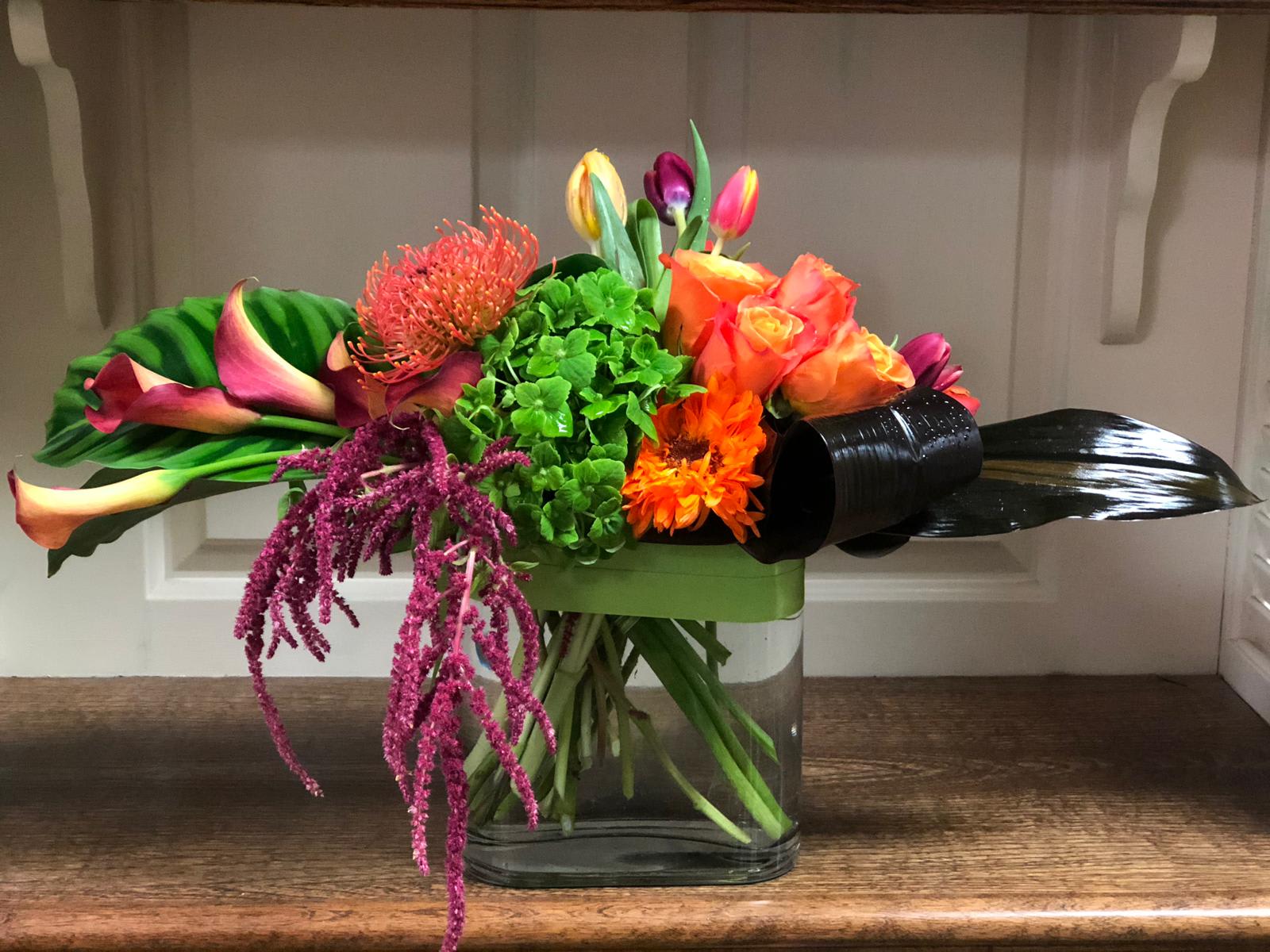 Bertha Yanez Creation - Recipe: Mango colored Calla Lilies, Green Hydrangeas, Orange Roses, Pink Tulips, Fuji Mums, Orange Gerbera, Amaranthus, and Pincushion Protea in a 6” glass vase with green ribbon. Seasonal: All year round Design View: Front Facing View Photo shown: Standard 